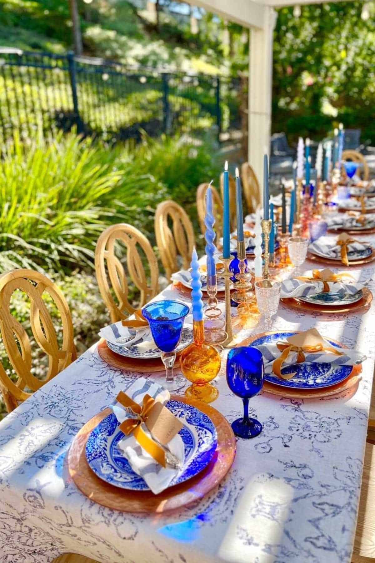 A long outdoor dining table is set with blue and gold glassware, patterned plates, gold flatware, and twisted candlesticks in various colors.