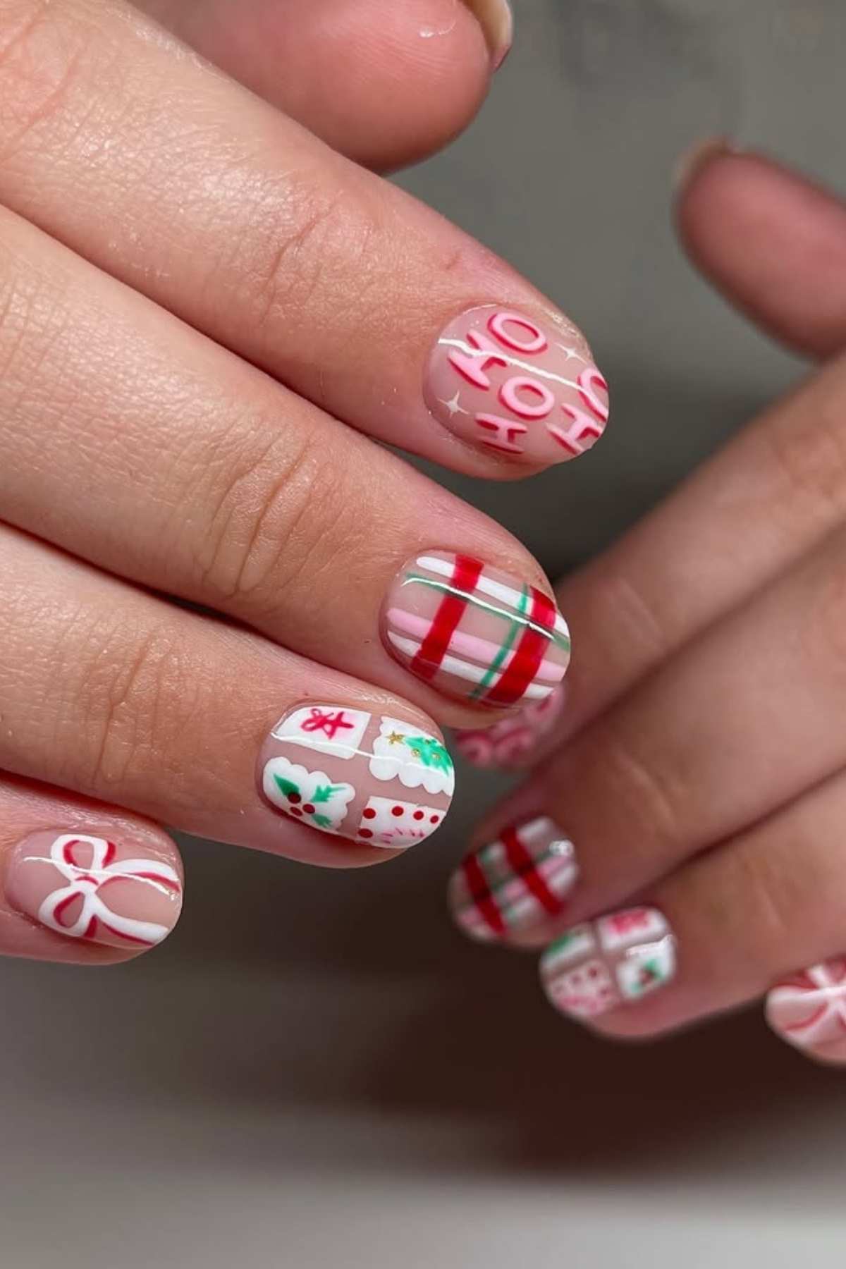 Close-up of hands with festive nail art featuring red, white, and green holiday designs such as stripes, bows, holly, and the words “Ho Ho Ho.