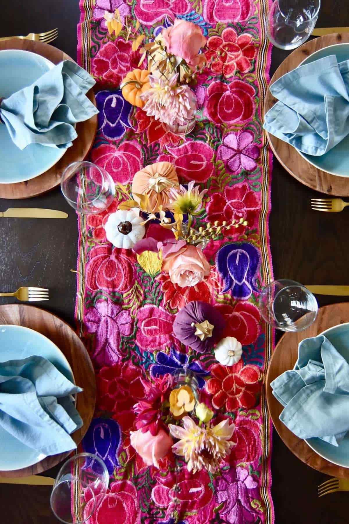 A table set with blue napkins, light plates on wooden chargers, gold cutlery, and a vibrant pink floral table runner with assorted flowers and small decorative pumpkins.