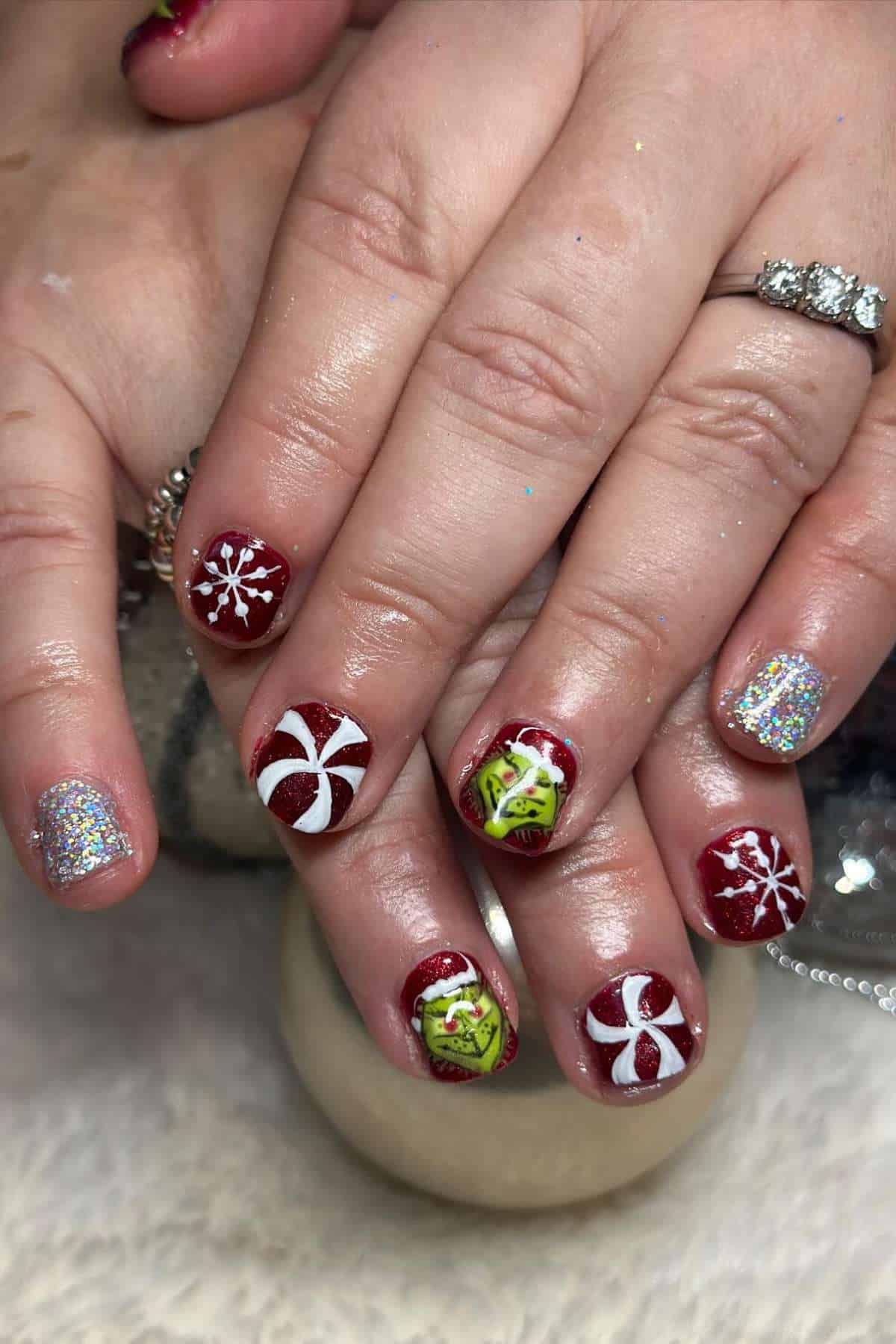 Hands with short, festive manicured nails featuring red polish, white snowflakes, peppermint designs, grinch's face, and accent nails with silver glitter.