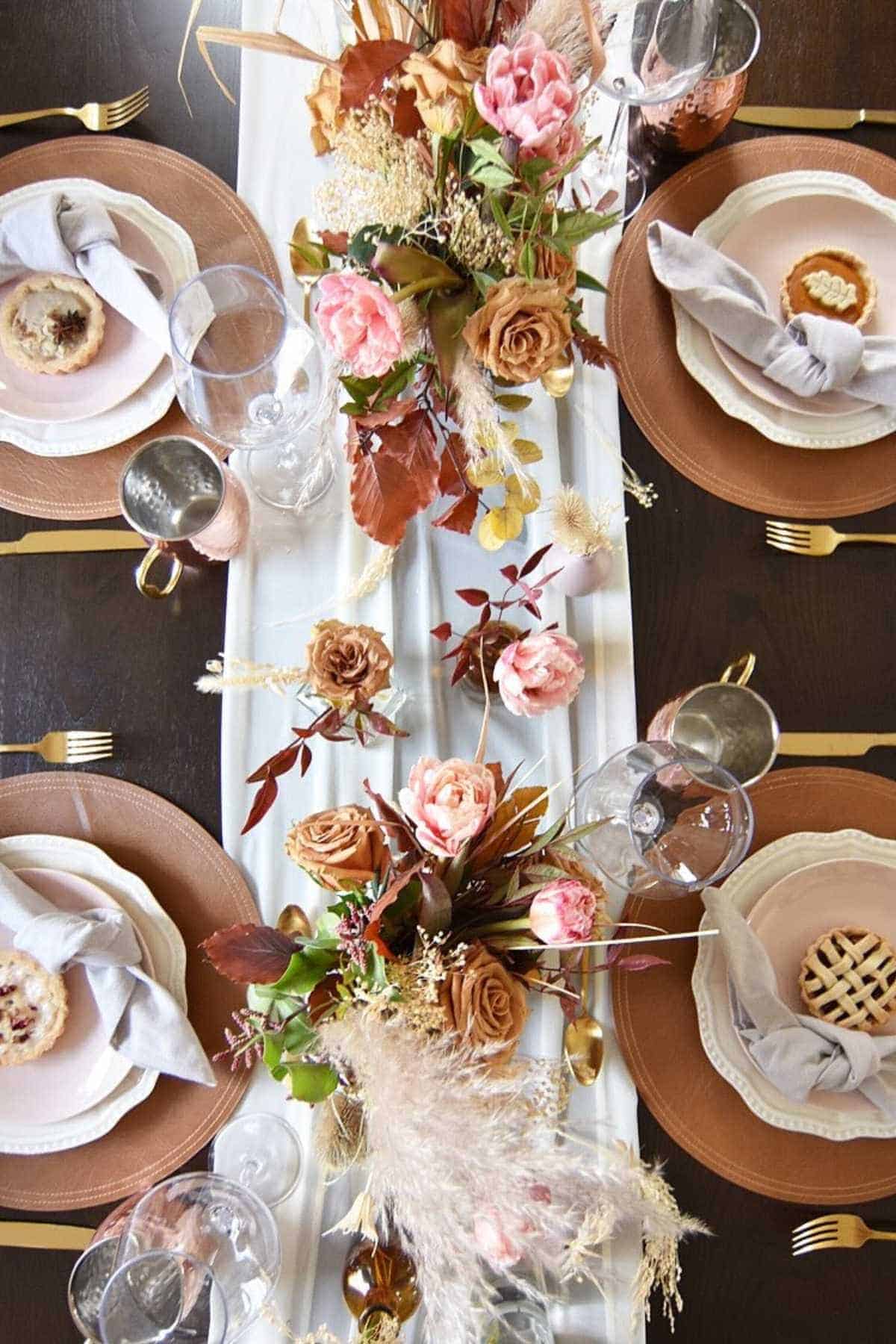 A dining table set for four with pink and tan floral centerpieces, gold cutlery, light gray napkins, and small individual pies on brown plates.