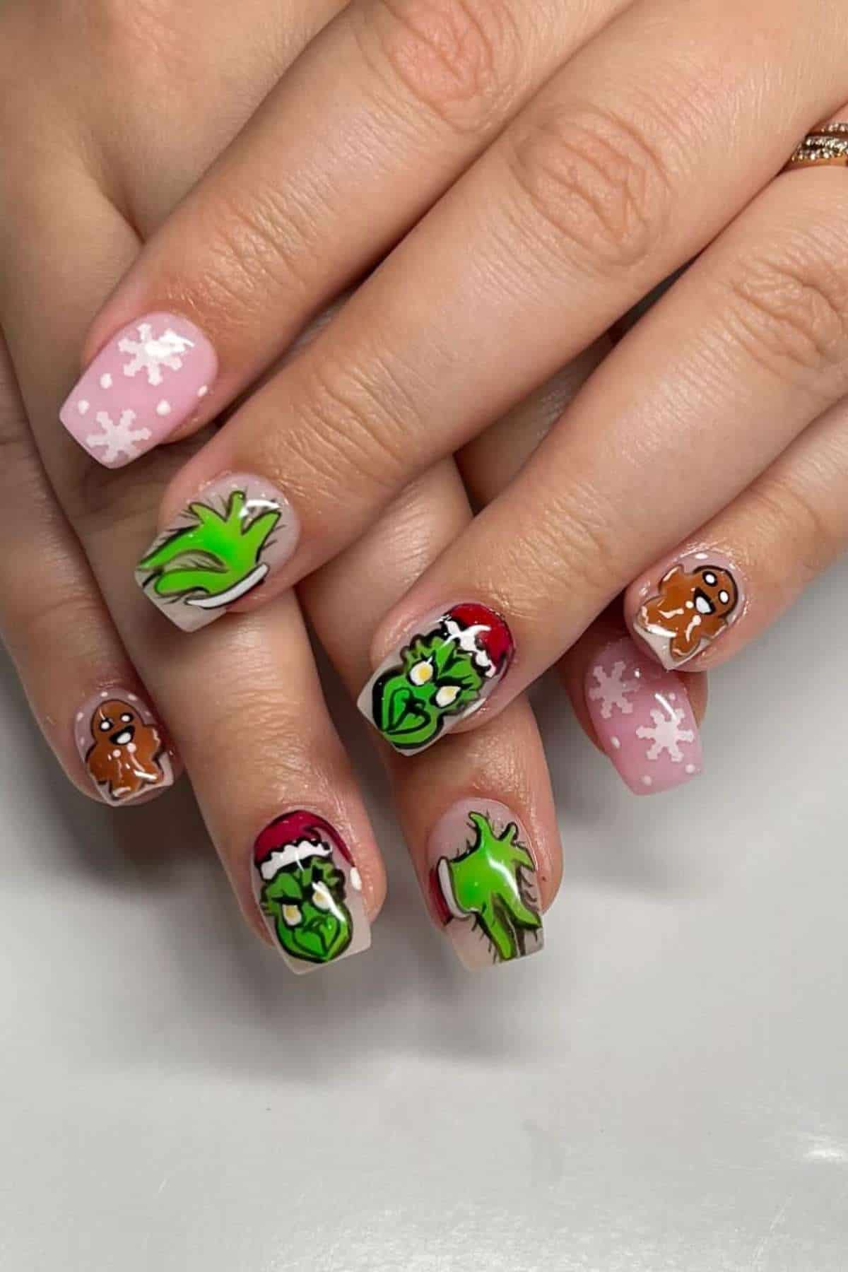 Close-up of hands with pink Christmas-themed nail art featuring snowflakes, gingerbread men, and the green character from "The Grinch" wearing a Santa hat on accent nails.