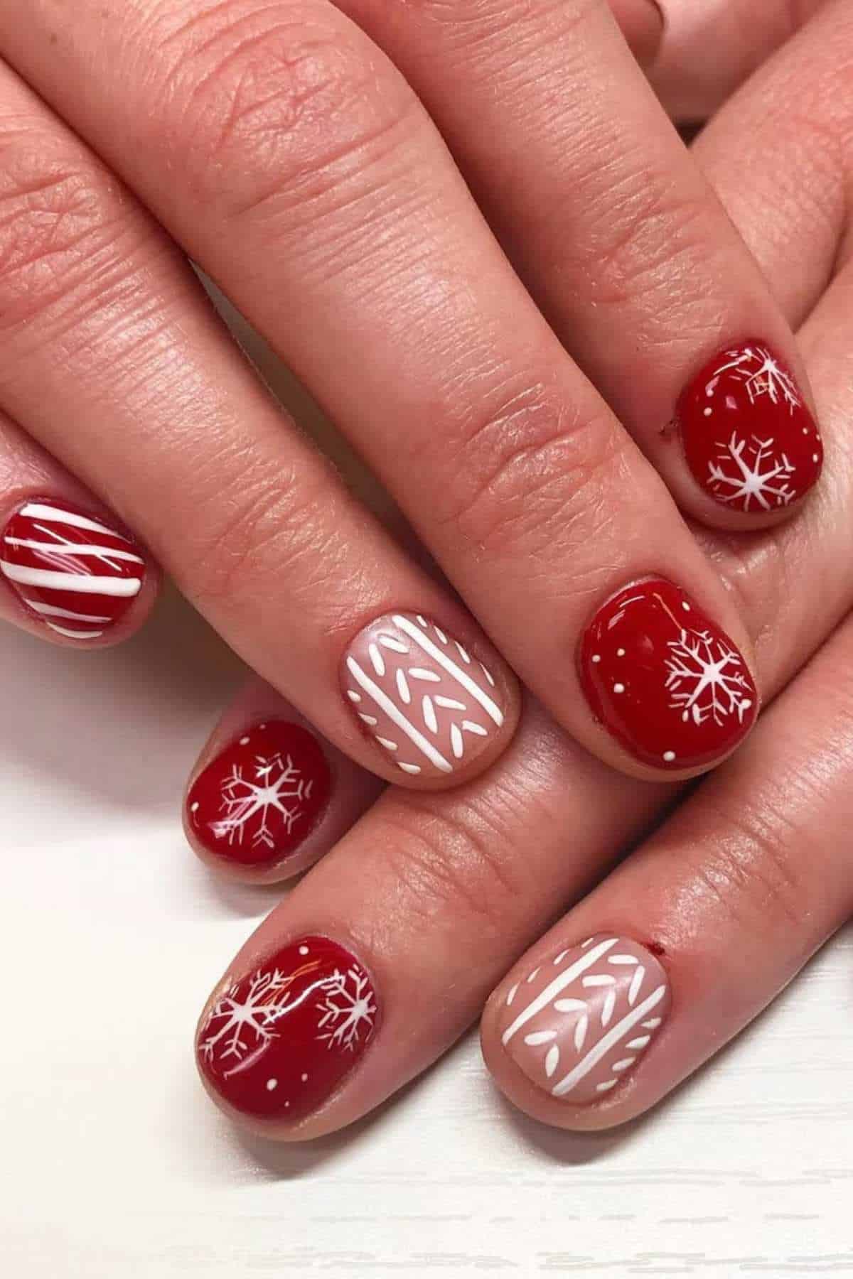 Hands with short nails painted in red and nude polish, featuring white snowflake and geometric designs for a festive look.