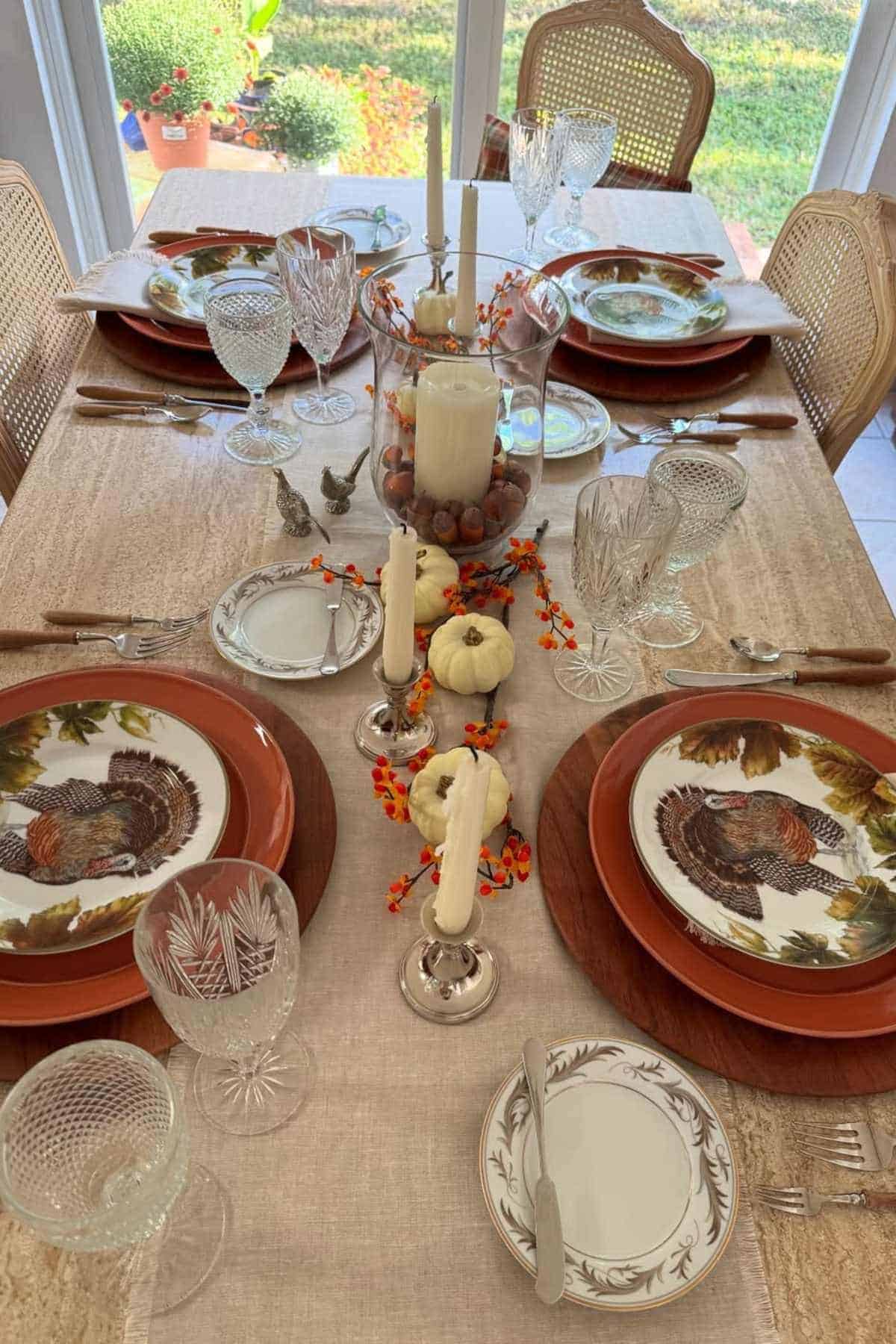 A dining table set for four with autumn-themed plates featuring turkeys, crystal glassware, candles, mini pumpkins, and orange berries as decoration.