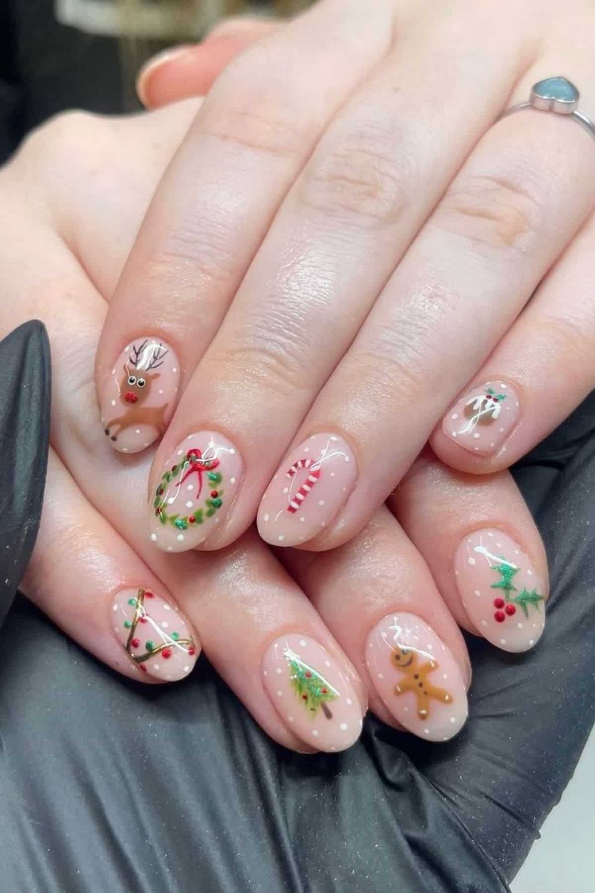A hand with Christmas-themed nail art, including designs of a reindeer, wreath, candy cane, holly, tree, gingerbread man, and lights on a nude base.