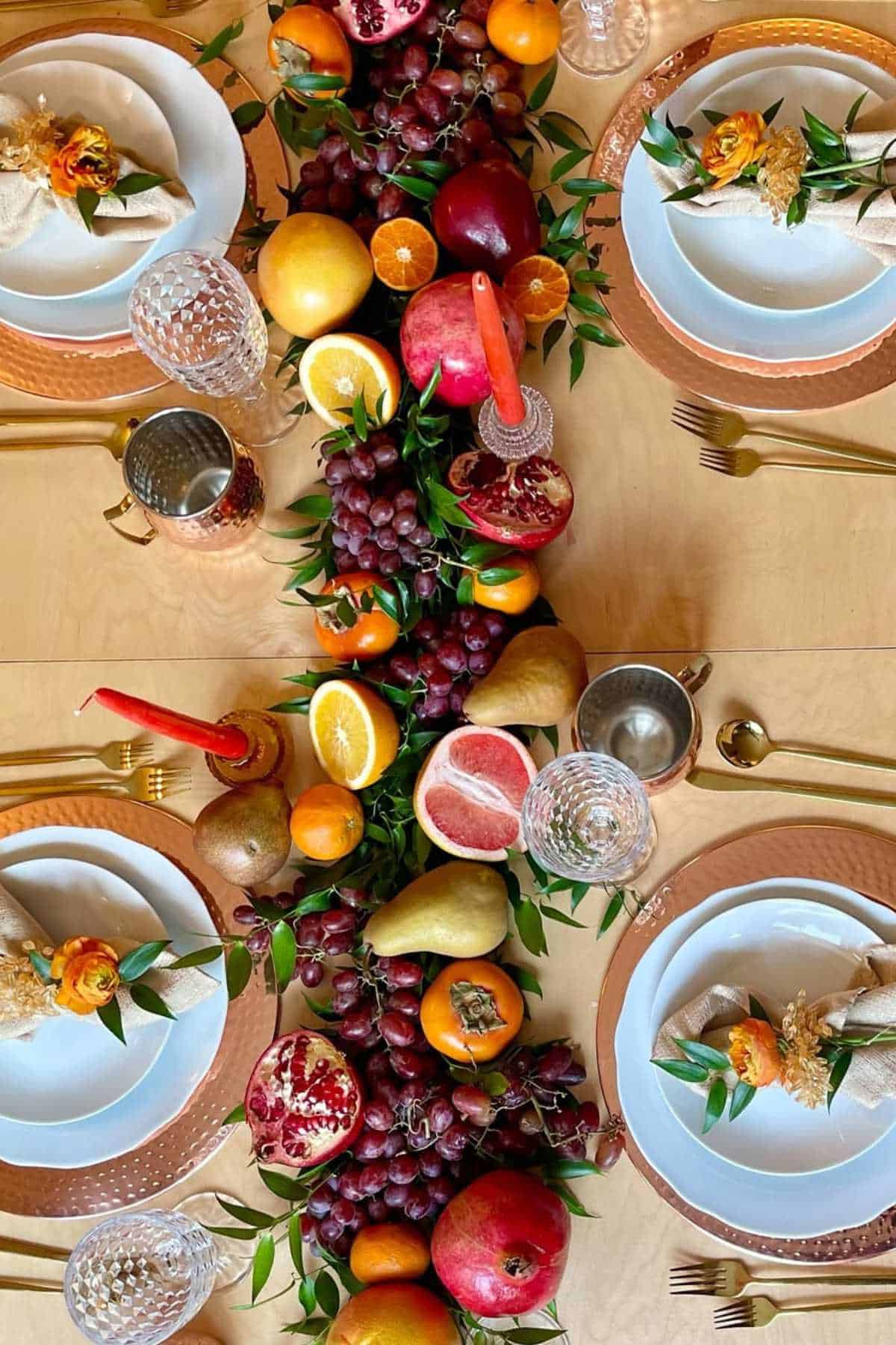 A table set with four place settings and a centerpiece of assorted fruits, including grapes, pomegranate, pears, oranges, and greenery, with gold cutlery and glassware.