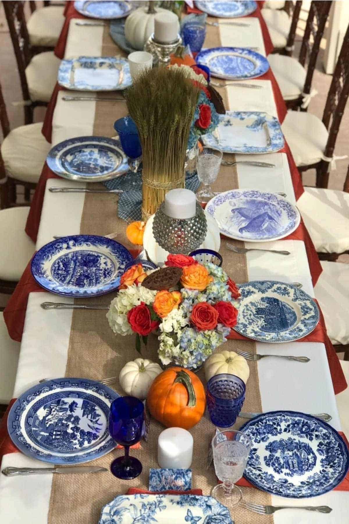 A dining table set with blue and white plates, blue glasses, pumpkins, floral arrangements, and wheat centerpieces on a burlap runner.