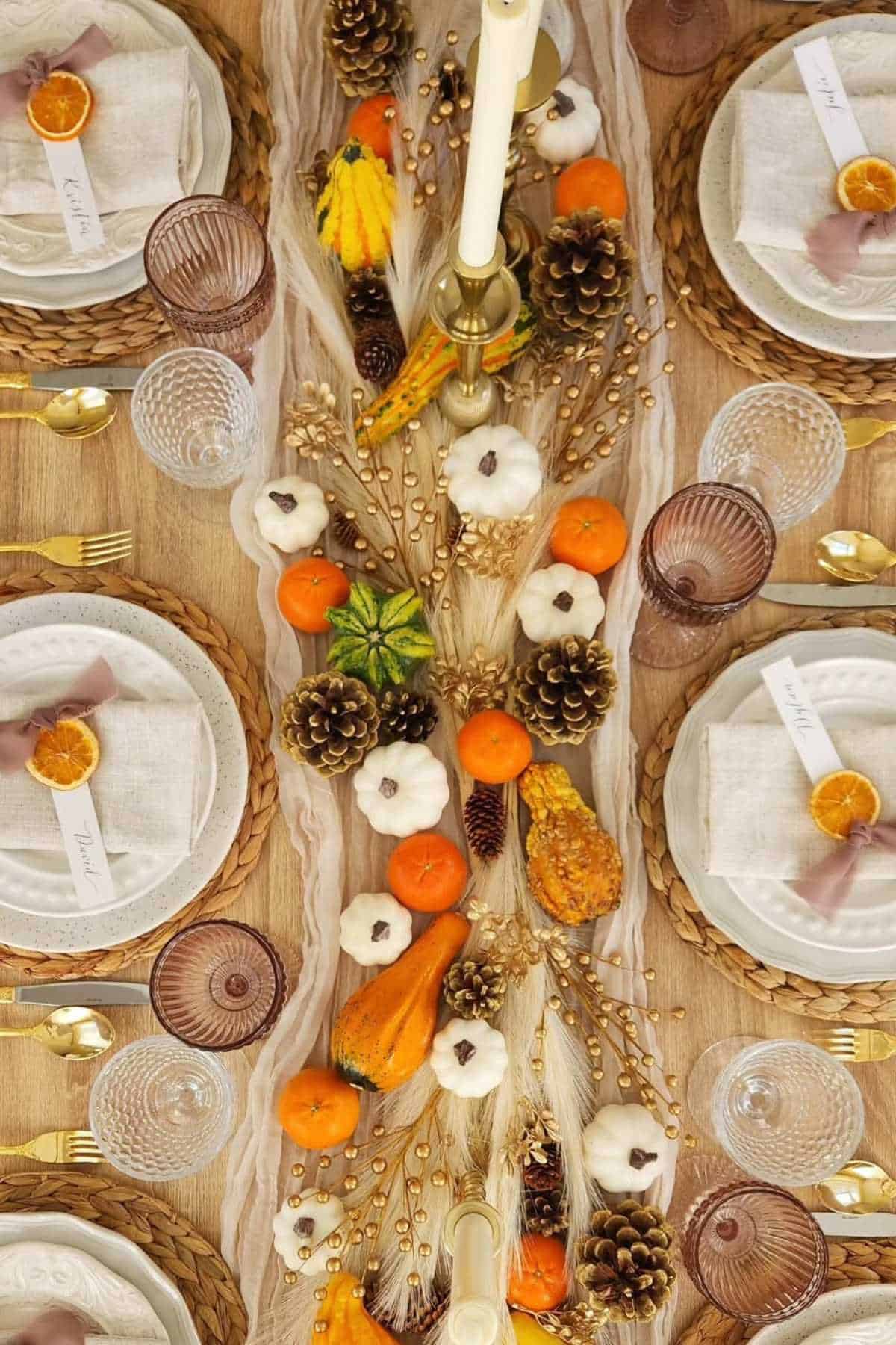 A fall-themed table setting with woven placemats, plates, gold cutlery, dried orange slices, mini pumpkins, gourds, pinecones, and decorative foliage arranged along the center.