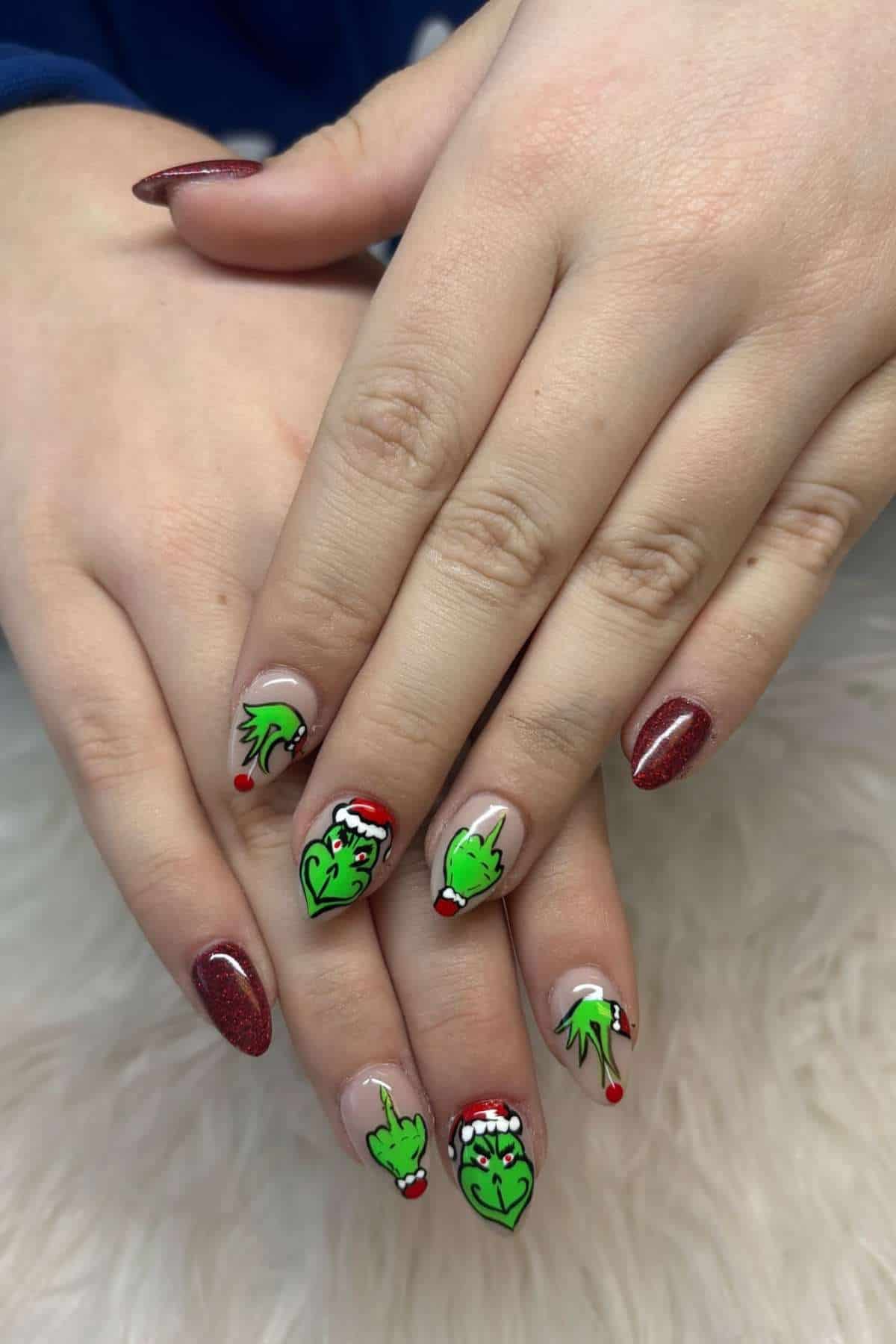 Hands with long nails featuring red glitter polish and festive Grinch-themed nail art, including green Grinch faces and Santa hats, displayed on a white textured surface.