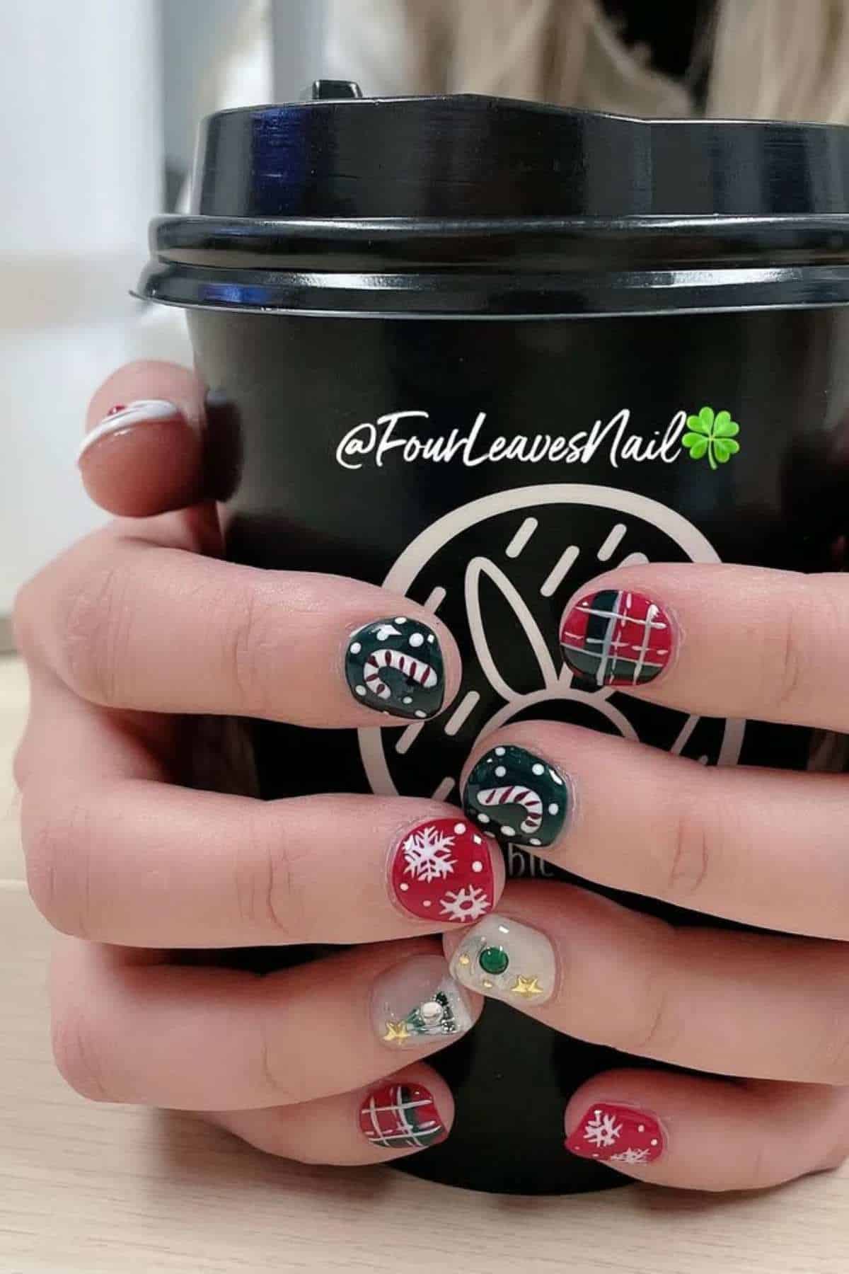 Close-up of hands holding a black coffee cup, featuring festive nail art with red, green, plaid patterns, snowflakes, and small holiday-themed designs.