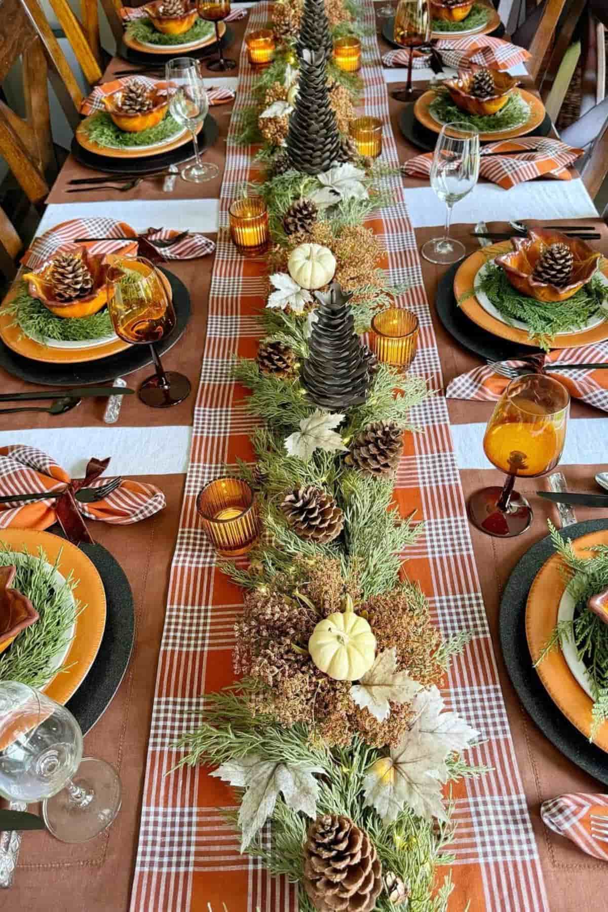 A long dining table with a plaid tablecloth, autumn-themed centerpiece of pinecones, small pumpkins, greenery, and brown table settings with orange and green accents.