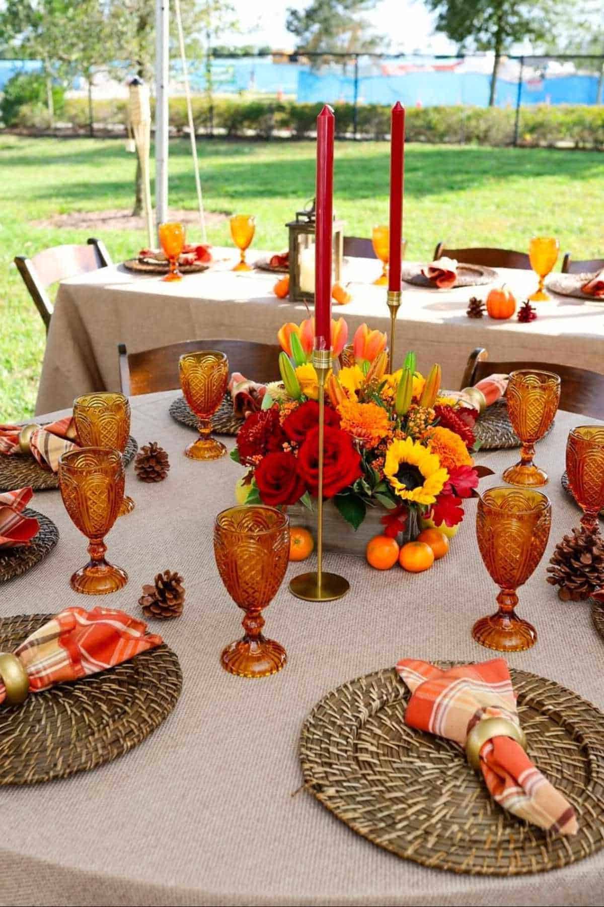 A round outdoor table is set with amber glassware, plaid napkins, woven placemats, pinecones, and a fall floral centerpiece with red candles.