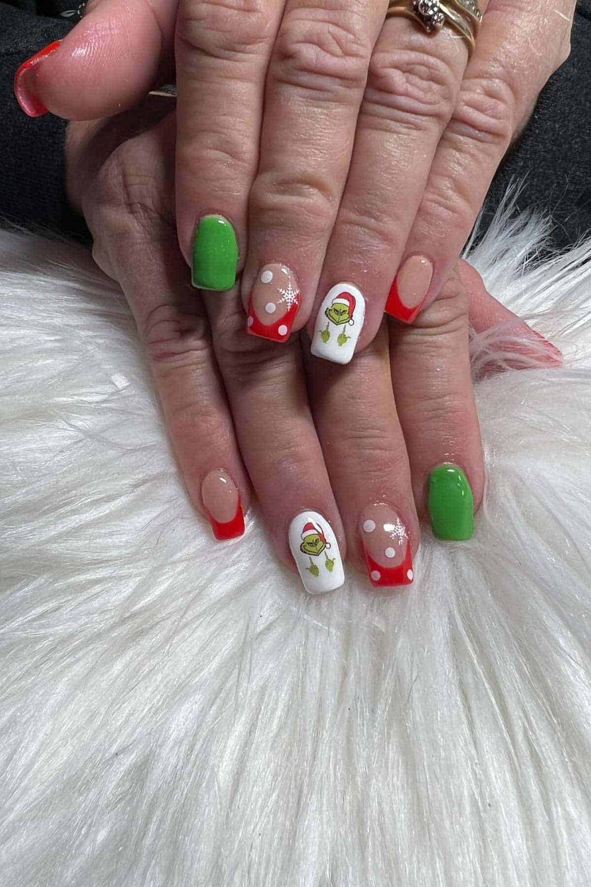 Hands with manicured nails featuring green and red polish, snowflake designs, and the Grinch illustrations, resting on a white furry surface.