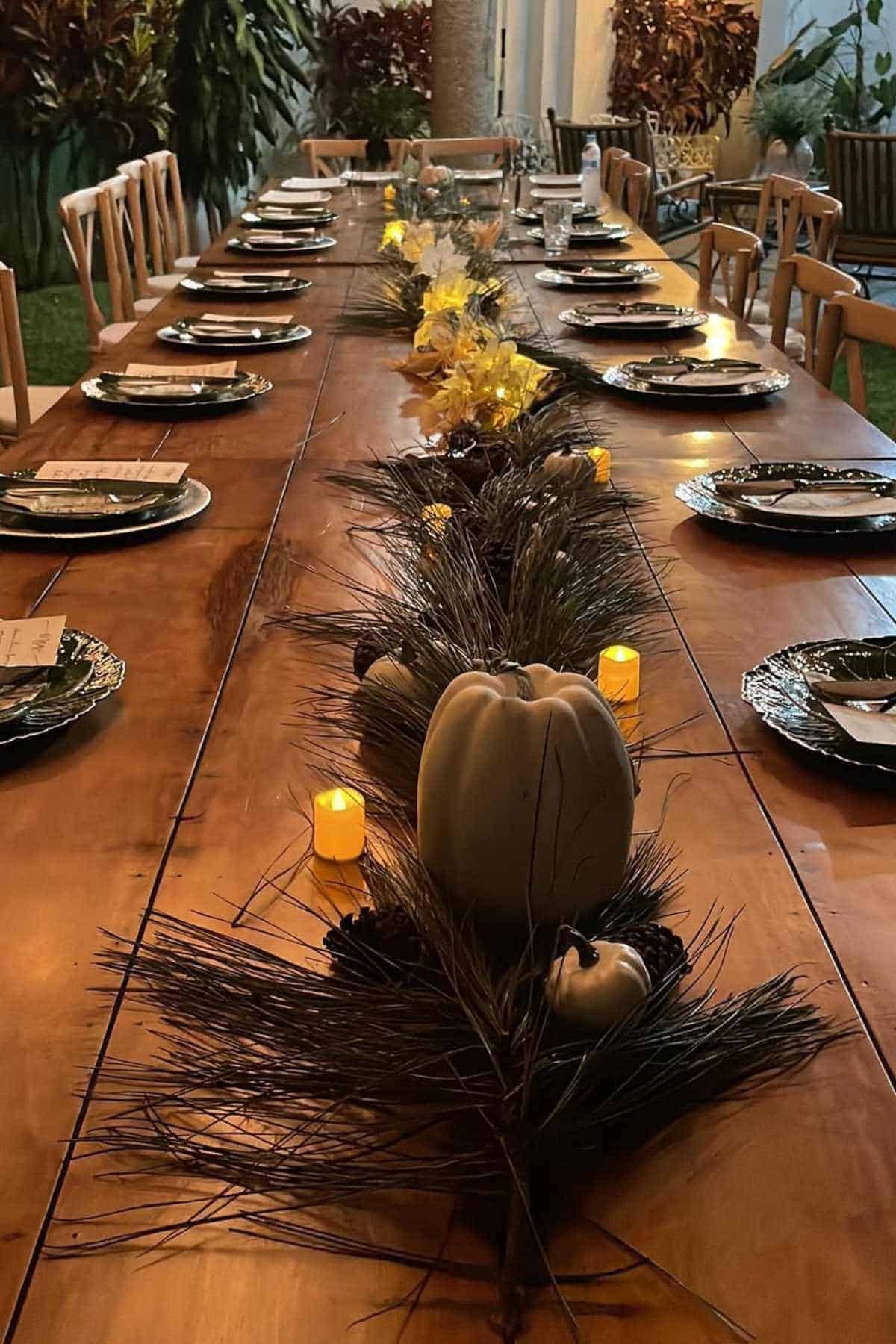 A long wooden dining table set with plates, napkins, and name cards, decorated with small pumpkins, pine branches, and lit candles as a centerpiece.