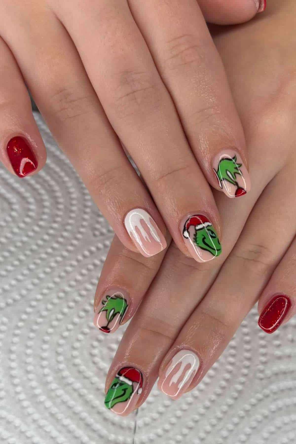 Hands with manicured nails featuring red polish, white designs, and green Grinch faces in Santa hats on a textured white surface.