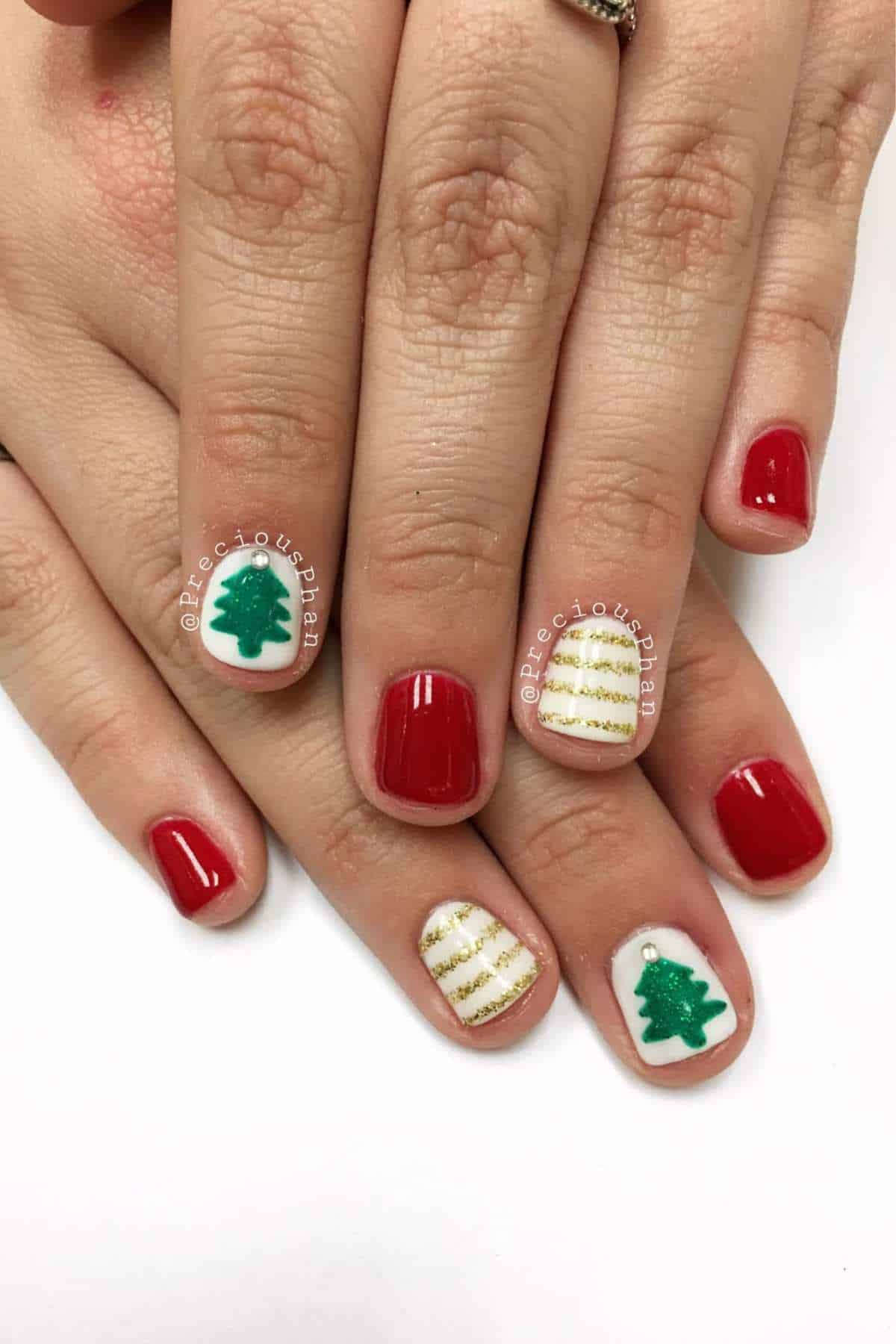 Hands with short nails painted red, except for two accent nails with white polish featuring a green Christmas tree and gold-striped design.
