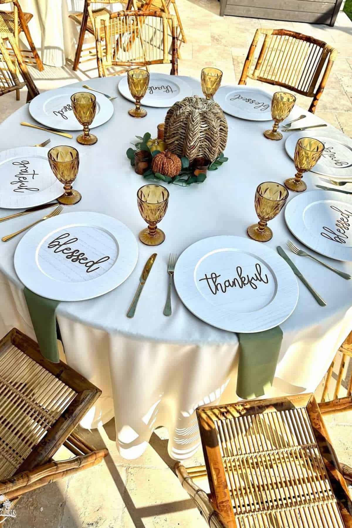 A round table set with woven chairs, amber glasses, white plates with words like "thankful" and "blessed," gold flatware, and a centerpiece of pumpkins and a turkey.