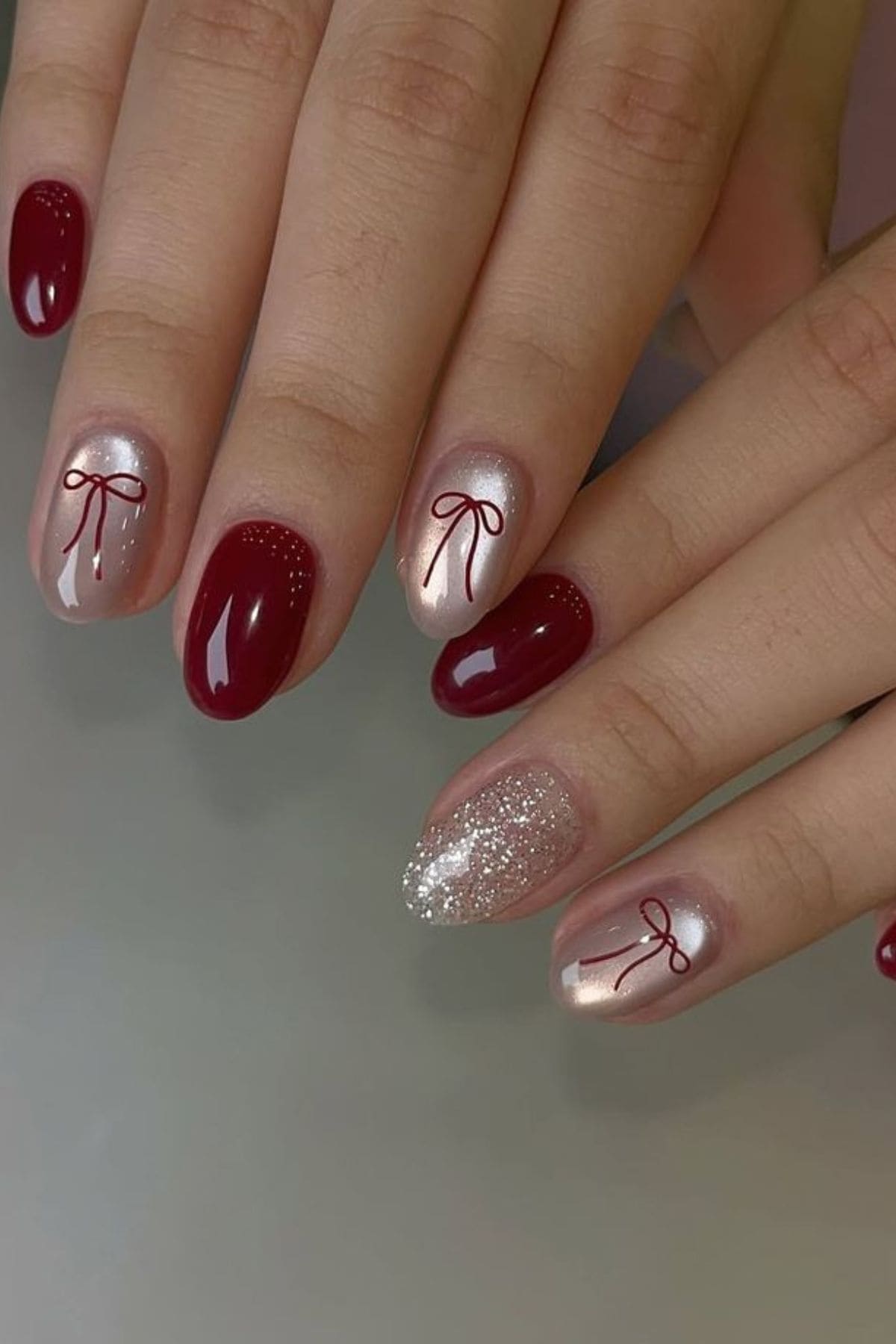 Two hands with manicured nails featuring a mix of dark red polish, nude polish with small red bow designs, and one nail with silver glitter accent.