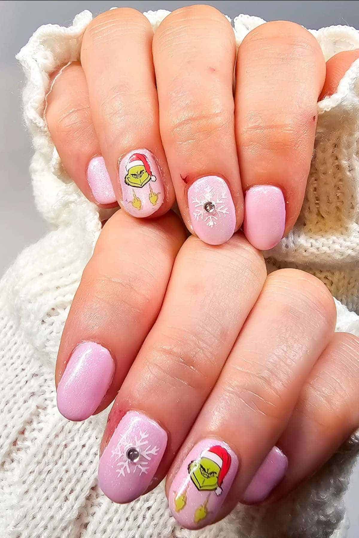 Hands with light pink, glossy nail polish featuring white snowflakes, rhinestone accents, and Grinch faces wearing Santa hats on two nails. Sweater sleeves are visible.