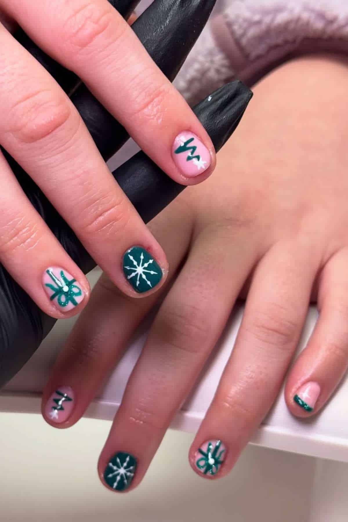 Hands with short nails painted dark green and light pink, featuring white and green holiday designs like snowflakes, stars, and abstract lines.
