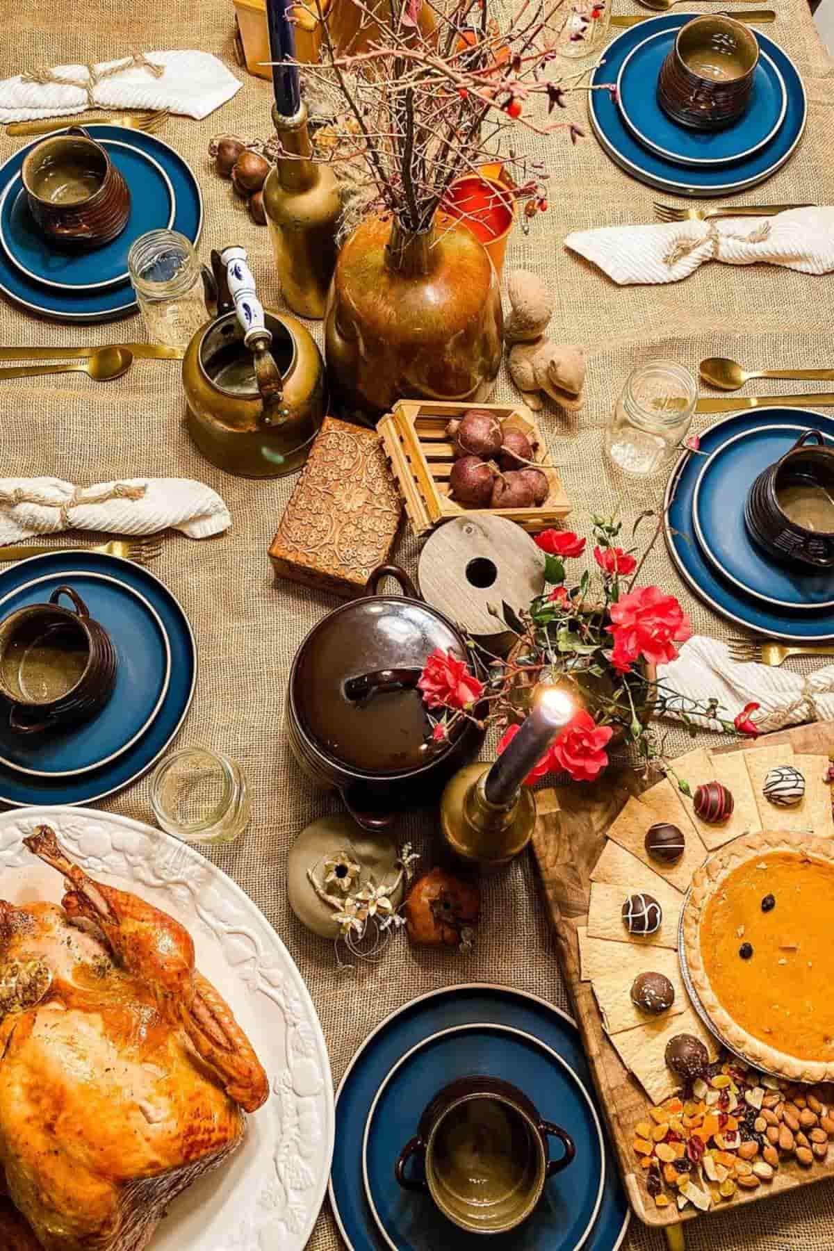 A festive dinner table set with blue plates, cups, a roast chicken, pumpkin pie, assorted chocolates, candles, and autumn-themed decorations.
