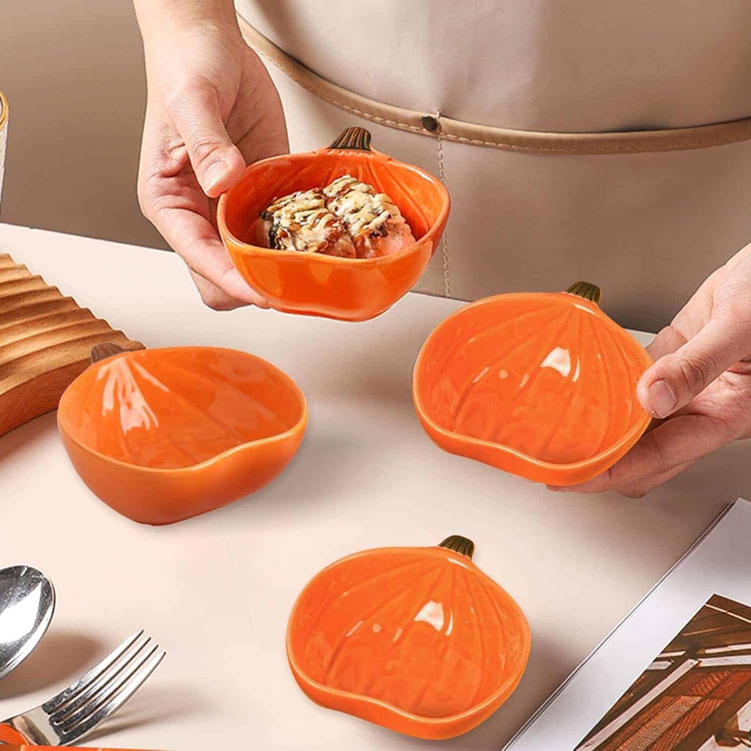A person holds a small orange pumpkin-shaped bowl with food inside, next to three empty matching bowls on a table set with utensils and a wooden tray.