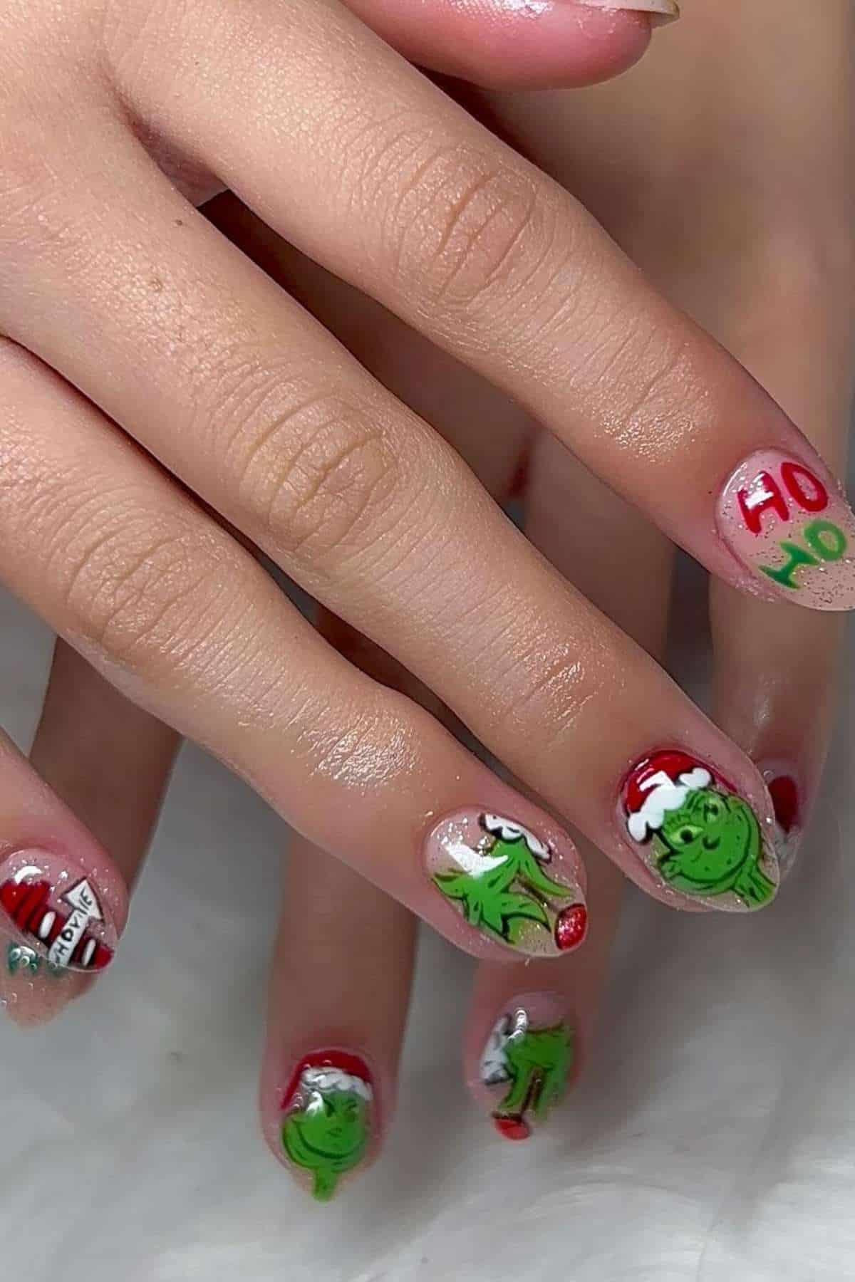 Close-up of hands with long fingernails painted with Christmas-themed designs, including green Grinch faces, Santa hats, "Ho Ho" text, and holiday decorations.