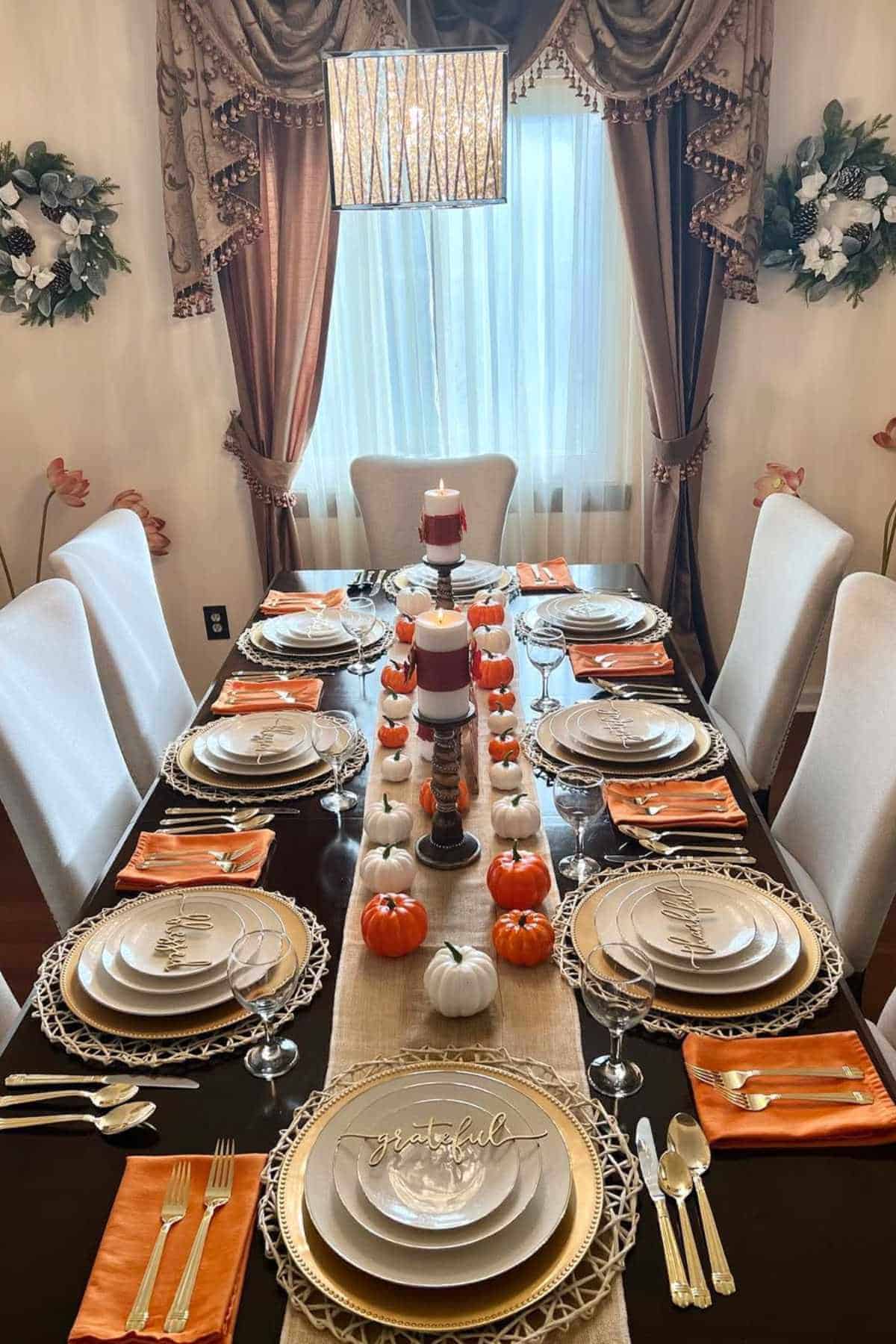 A formal dining table is set for eight, decorated with small pumpkins, stacked candles, and orange napkins, with natural light streaming through a curtained window.