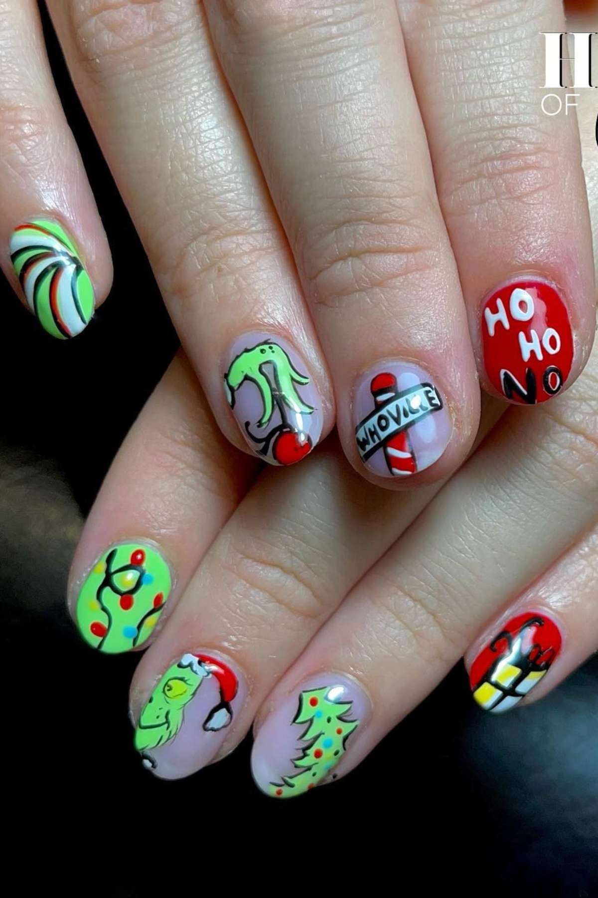 A hand with Christmas-themed nail art including designs of the Grinch, candy cane, Christmas tree, ornaments, "HO HO NO" text, and a "Whoville" sign.