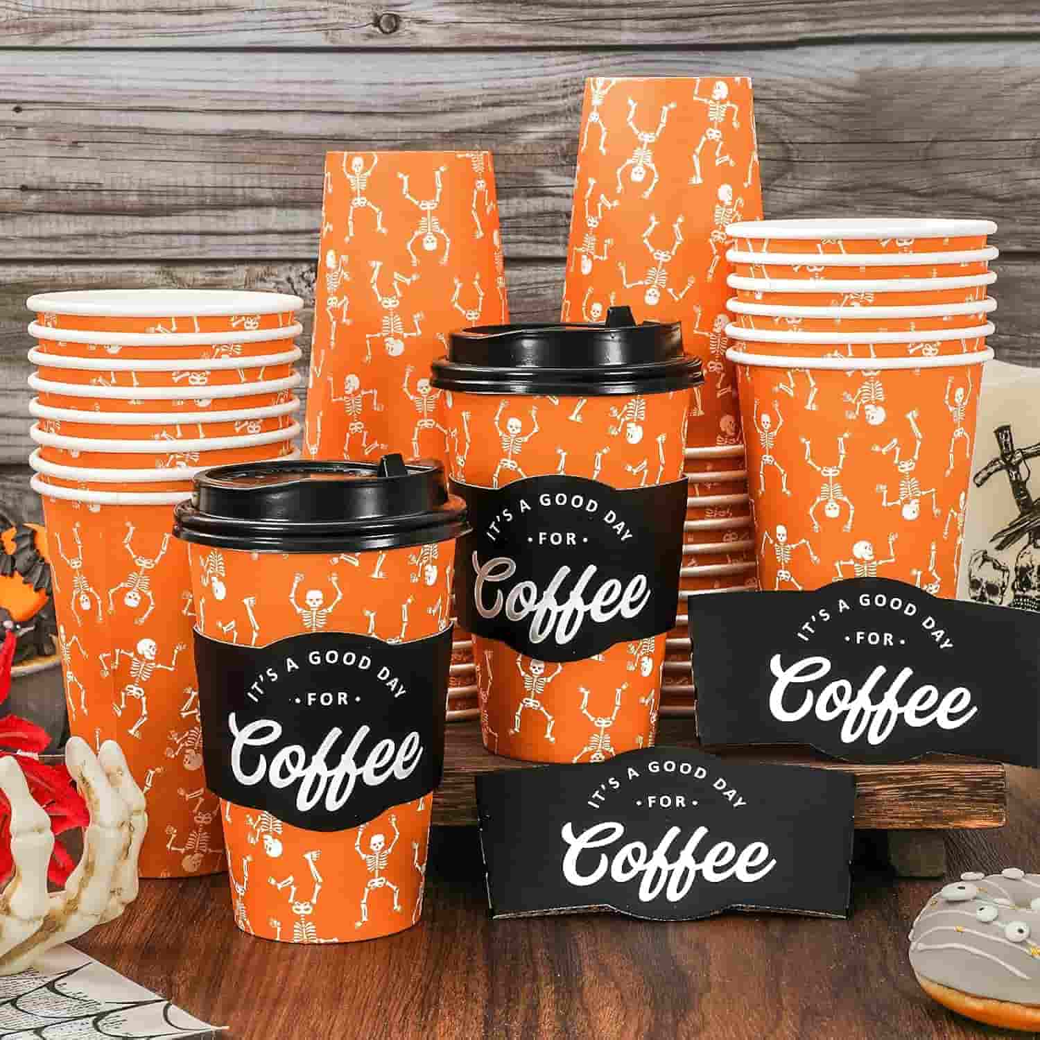 Several orange coffee cups with skeleton designs and black sleeves reading "It's a good day for coffee" are stacked on a wooden surface with Halloween decorations nearby.