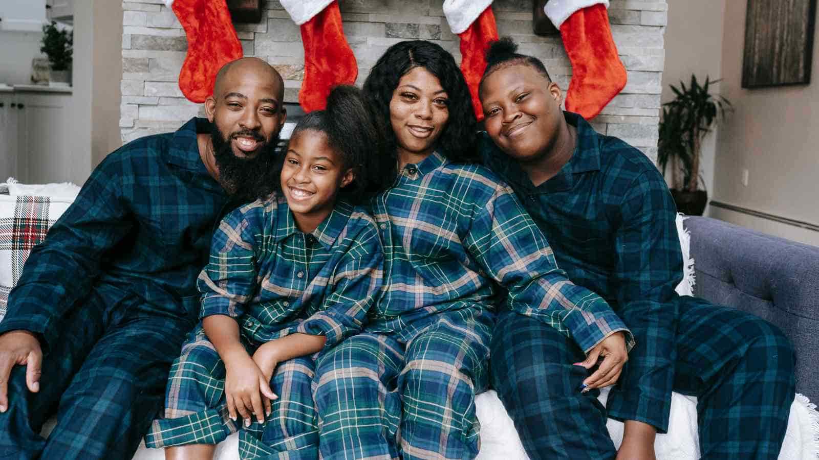 A family of four wearing matching plaid pajamas sits on a couch in front of a fireplace with red Christmas stockings hanging above them.