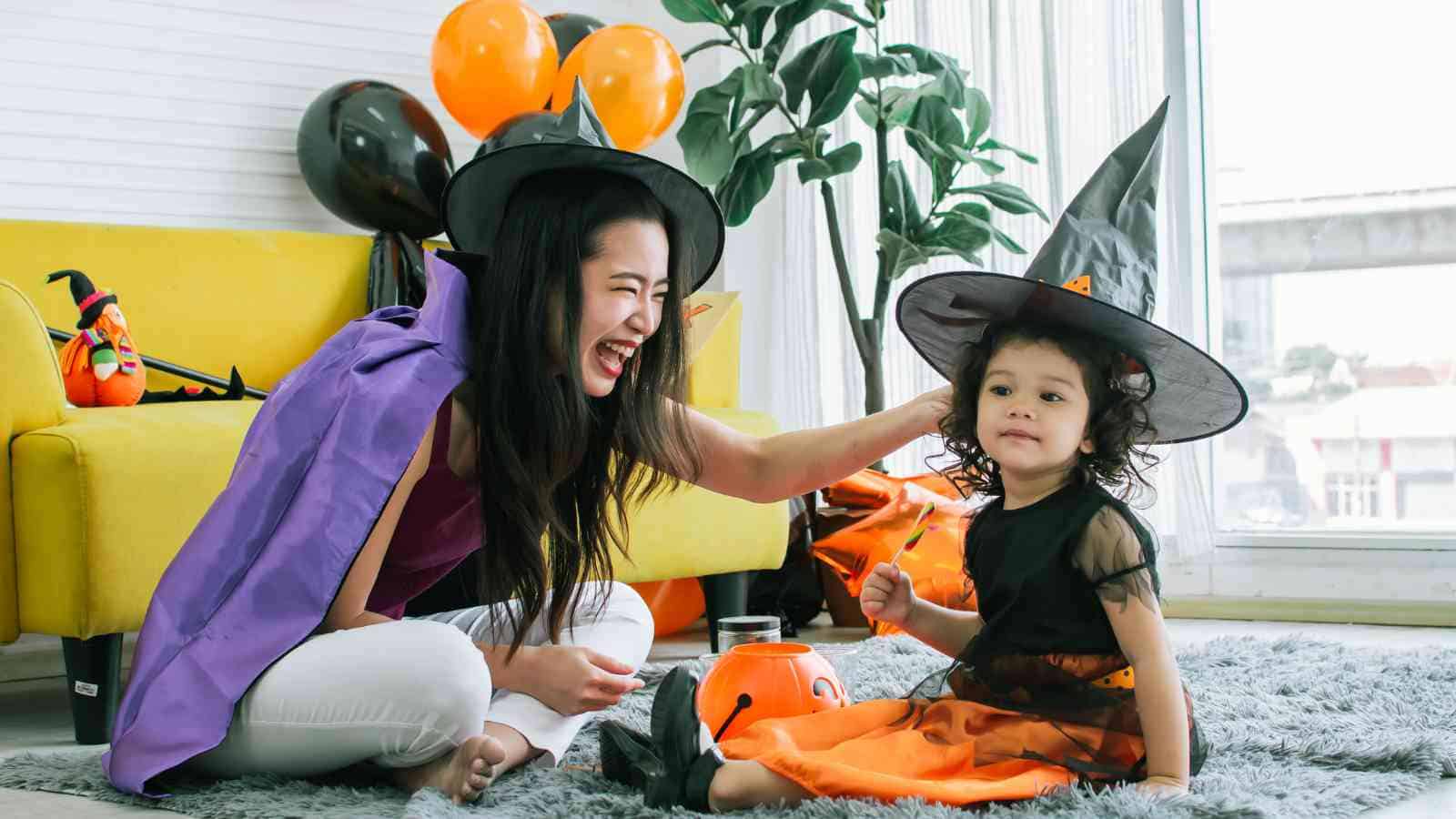An adult and a child in witch costumes sit on the floor, smiling, with Halloween decorations and balloons in the background.