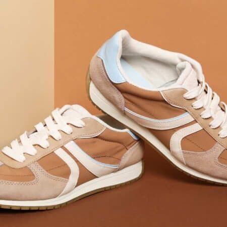 A pair of tan, beige, and white sneakers with white laces and light blue accents displayed on a brown surface against a beige background.