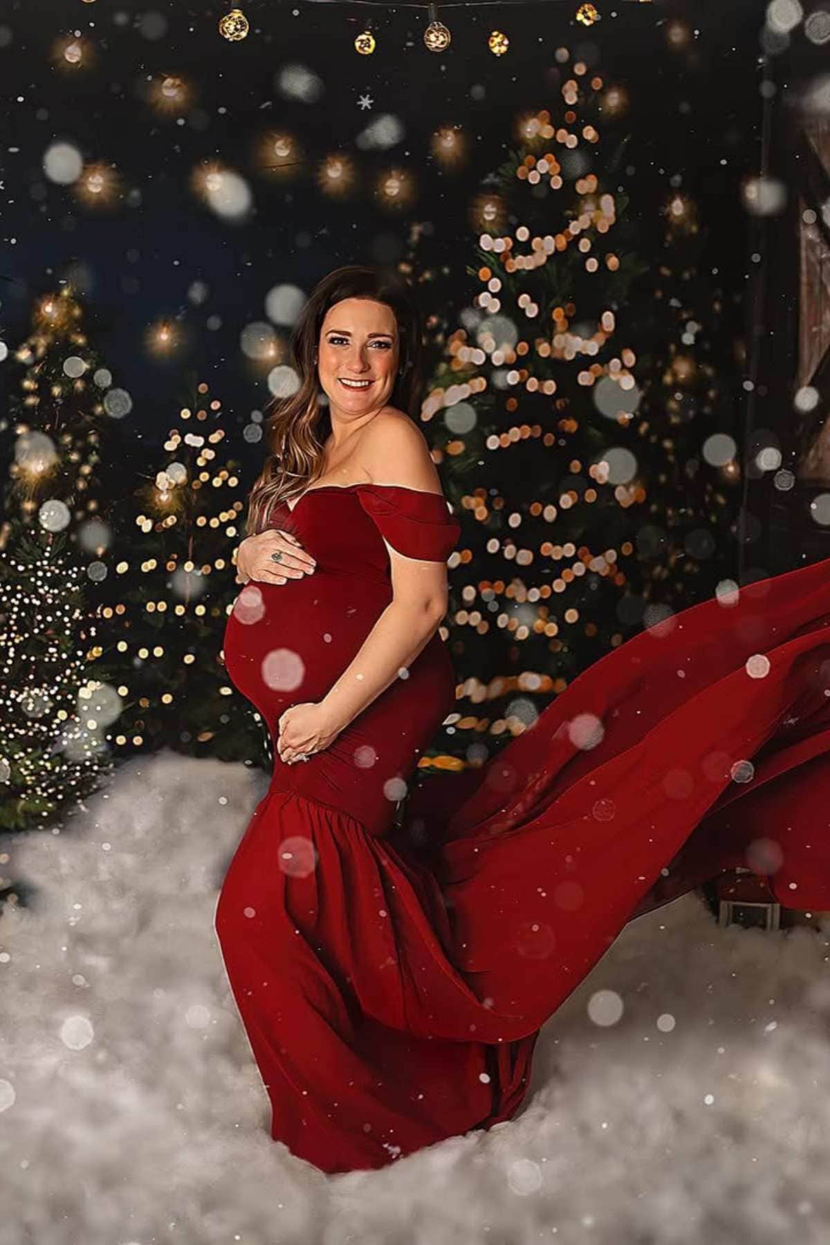 A pregnant woman in a red dress stands in a snowy, festive setting with decorated Christmas trees and string lights in the background.