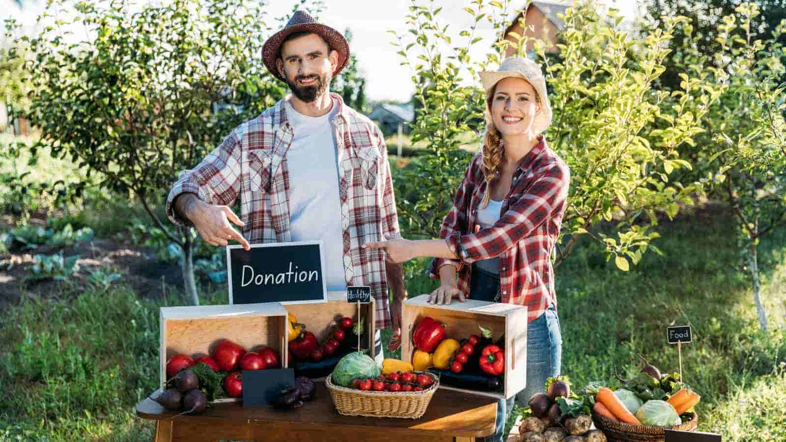 Two people stand at an outdoor table with fresh produce and a donation sign, smiling, surrounded by garden plants and trees.