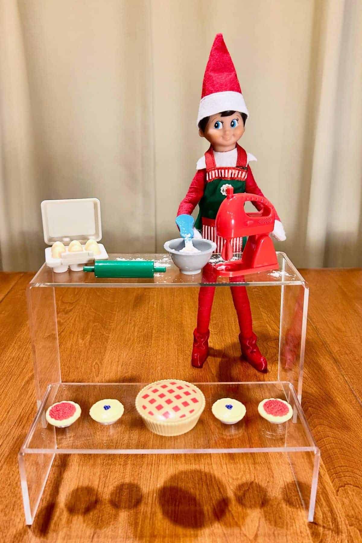 A toy elf dressed as a baker stands behind a mixing bowl and mixer, with mini pies and baking items displayed on a clear acrylic stand on a wooden table.