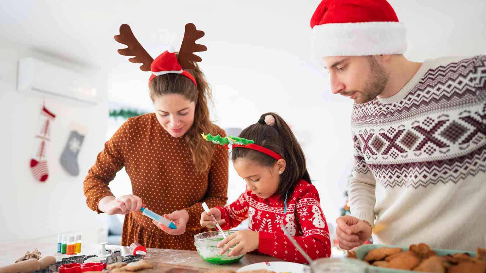Three people wearing festive sweaters and holiday headbands decorate cookies together in a bright kitchen, with Christmas stockings hanging in the background.