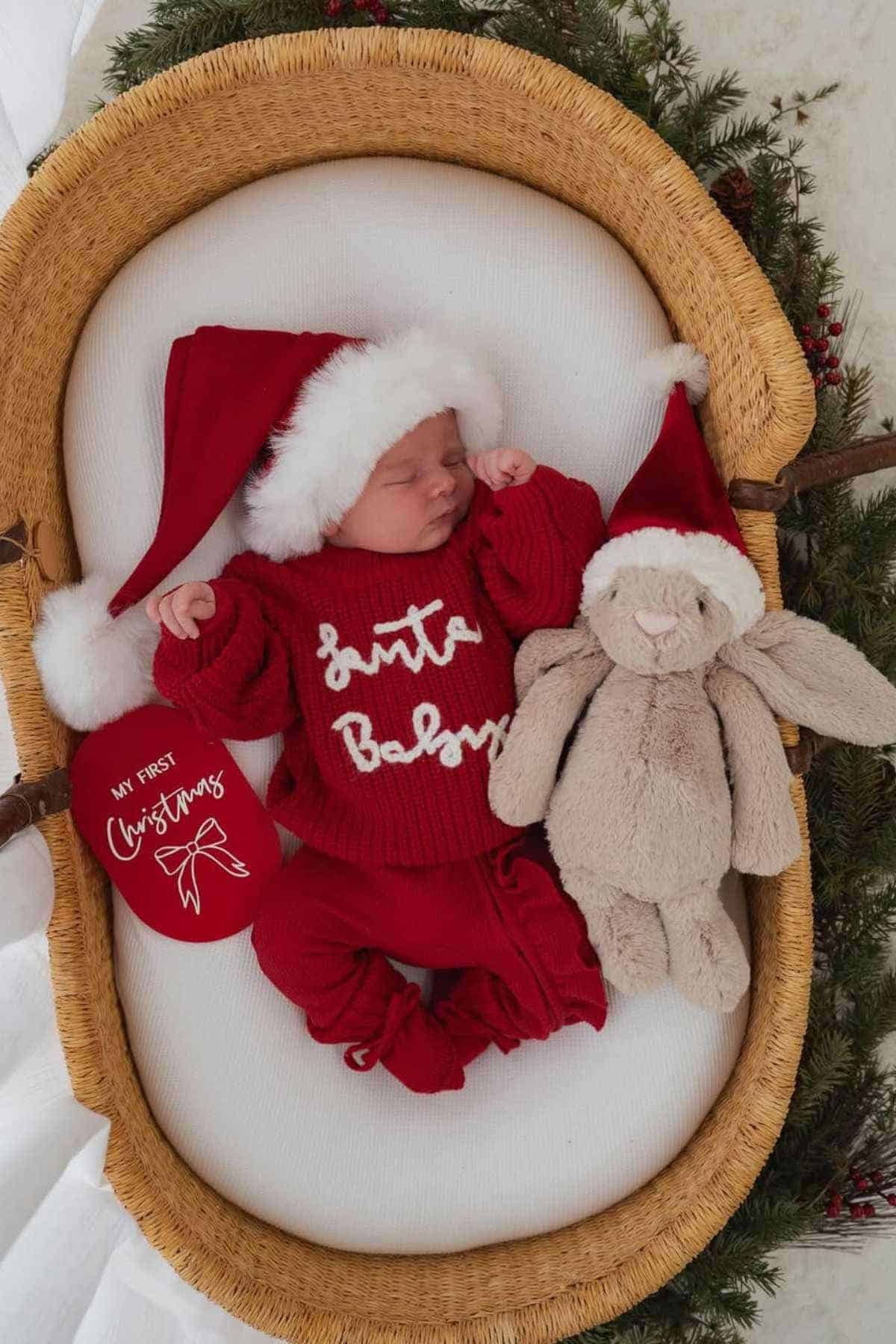 Newborn in a basket wearing a red "Santa Baby" outfit and Santa hat, lying beside a plush bunny and a "My First Christmas" ornament, surrounded by greenery.