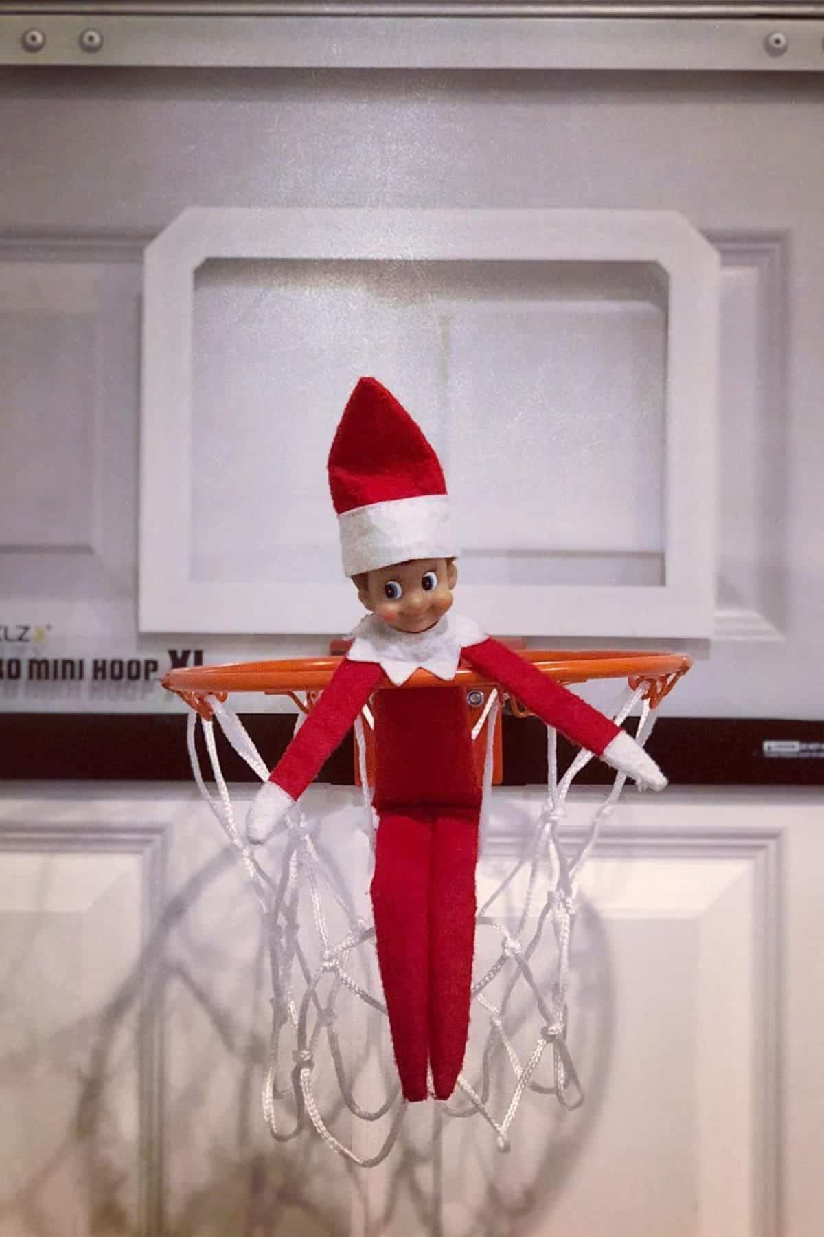 An Elf on the Shelf doll is sitting in the net of a small indoor basketball hoop attached to a door.