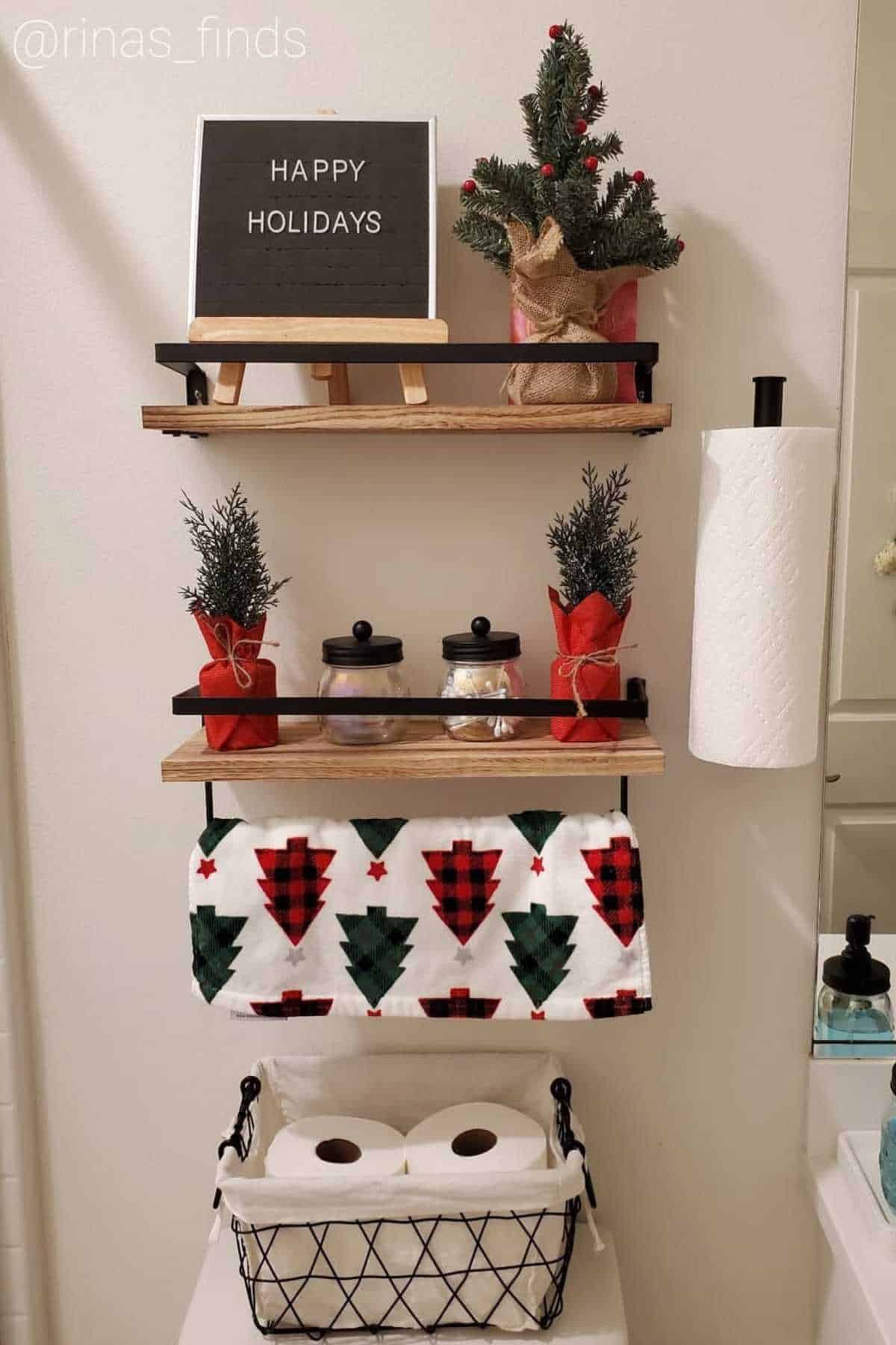 Bathroom shelves decorated for the holidays with small potted trees, a "Happy Holidays" sign, jars, a festive towel, and a basket holding toilet paper rolls.