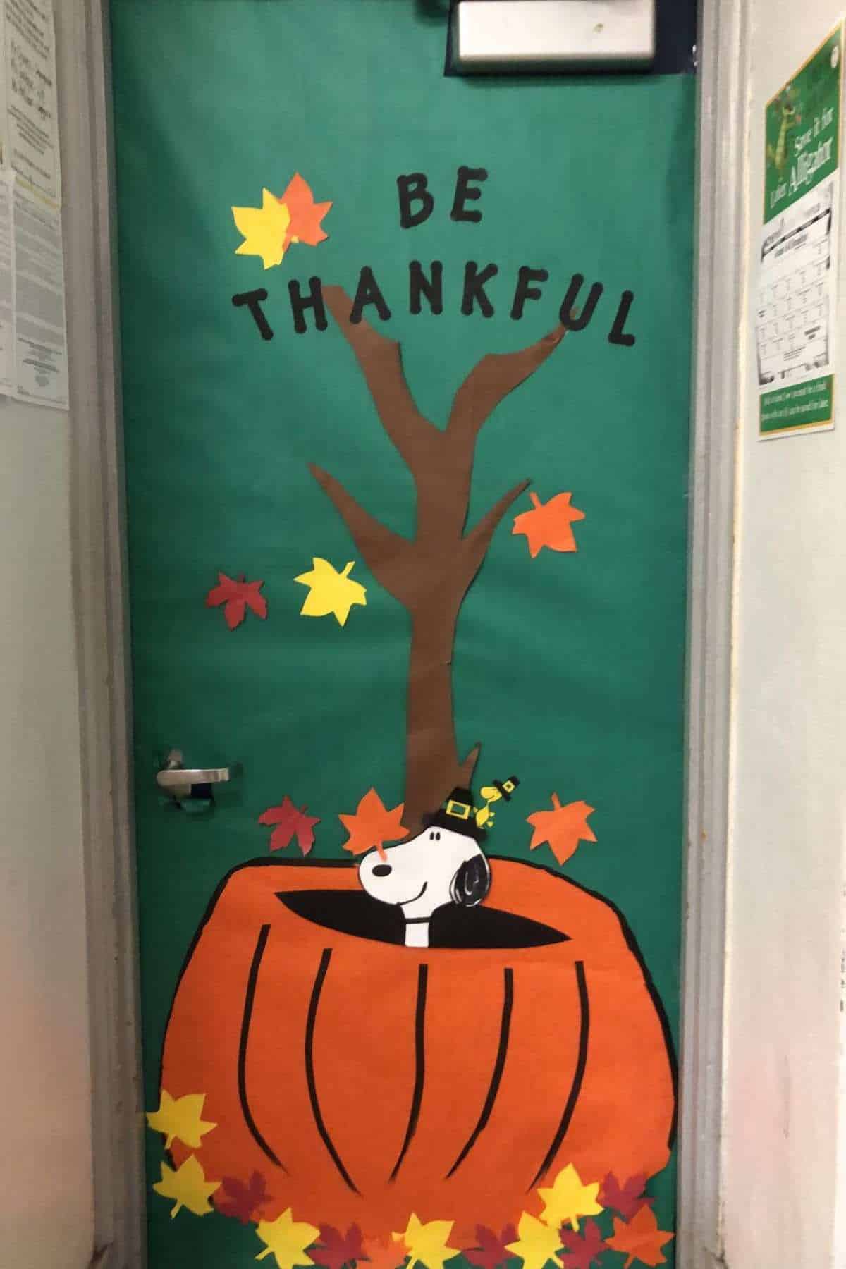 A decorated door features Snoopy in a pumpkin, a tree with autumn leaves, and the words "BE THANKFUL" on a green background.