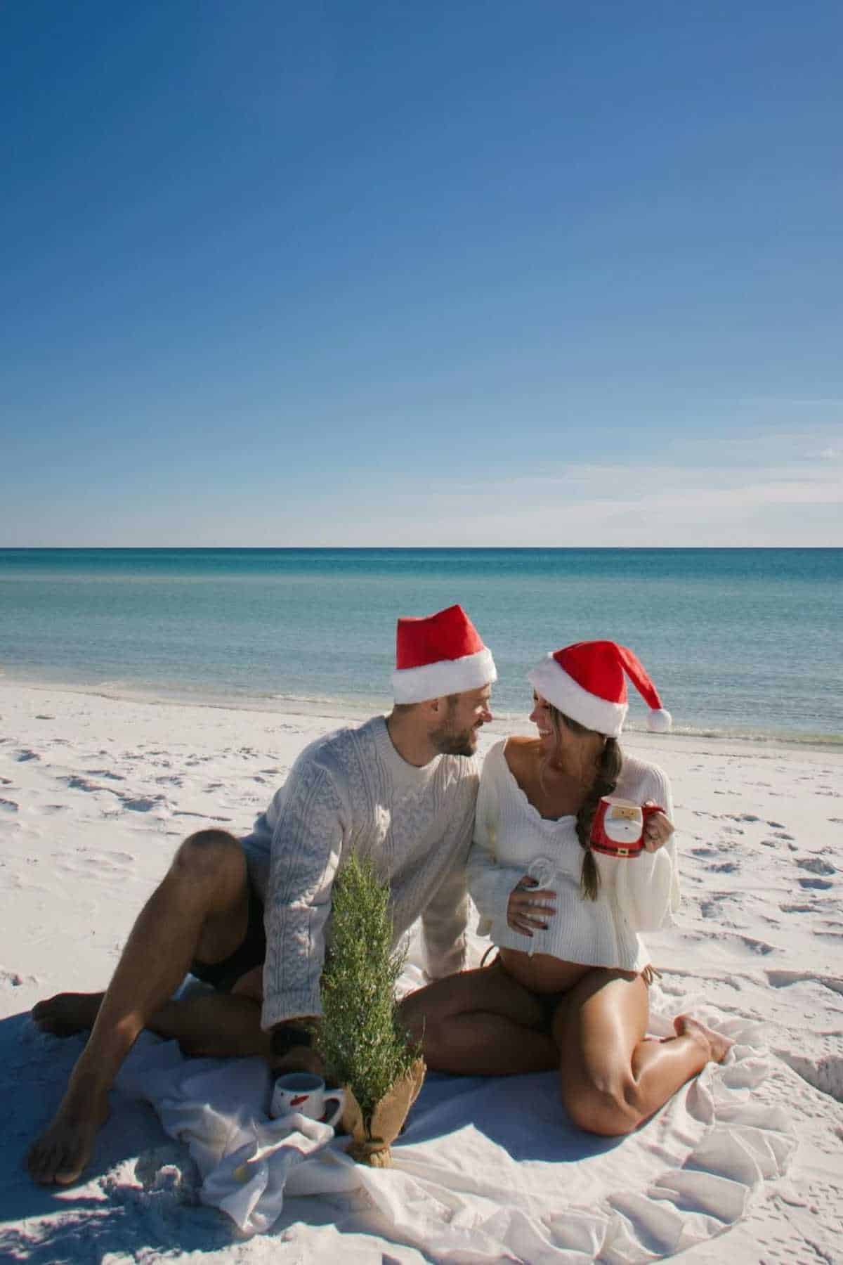 A couple wearing Santa hats sits on a beach blanket near a small Christmas tree, with clear blue sky and ocean in the background.
