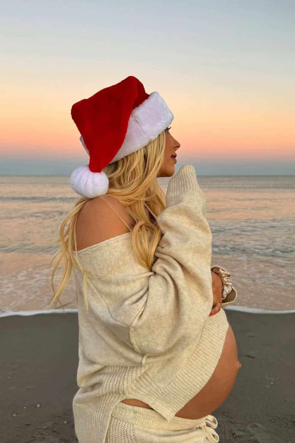 Pregnant woman in a Santa hat and beige sweater stands on the beach at sunset, looking toward the ocean.
