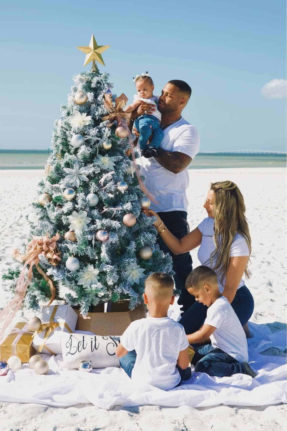 A family decorates a Christmas tree on a sandy beach with the ocean in the background; two adults, two young boys, and a baby are present.