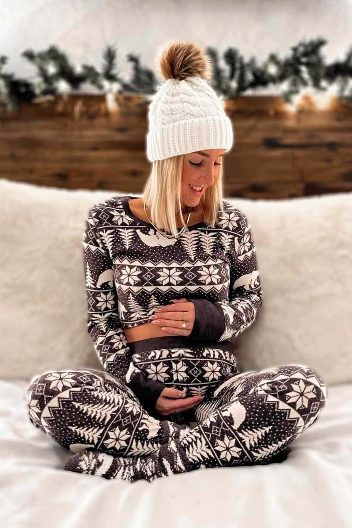 A woman in patterned winter pajamas and a white knit hat sits cross-legged on a bed, looking down and holding her stomach.