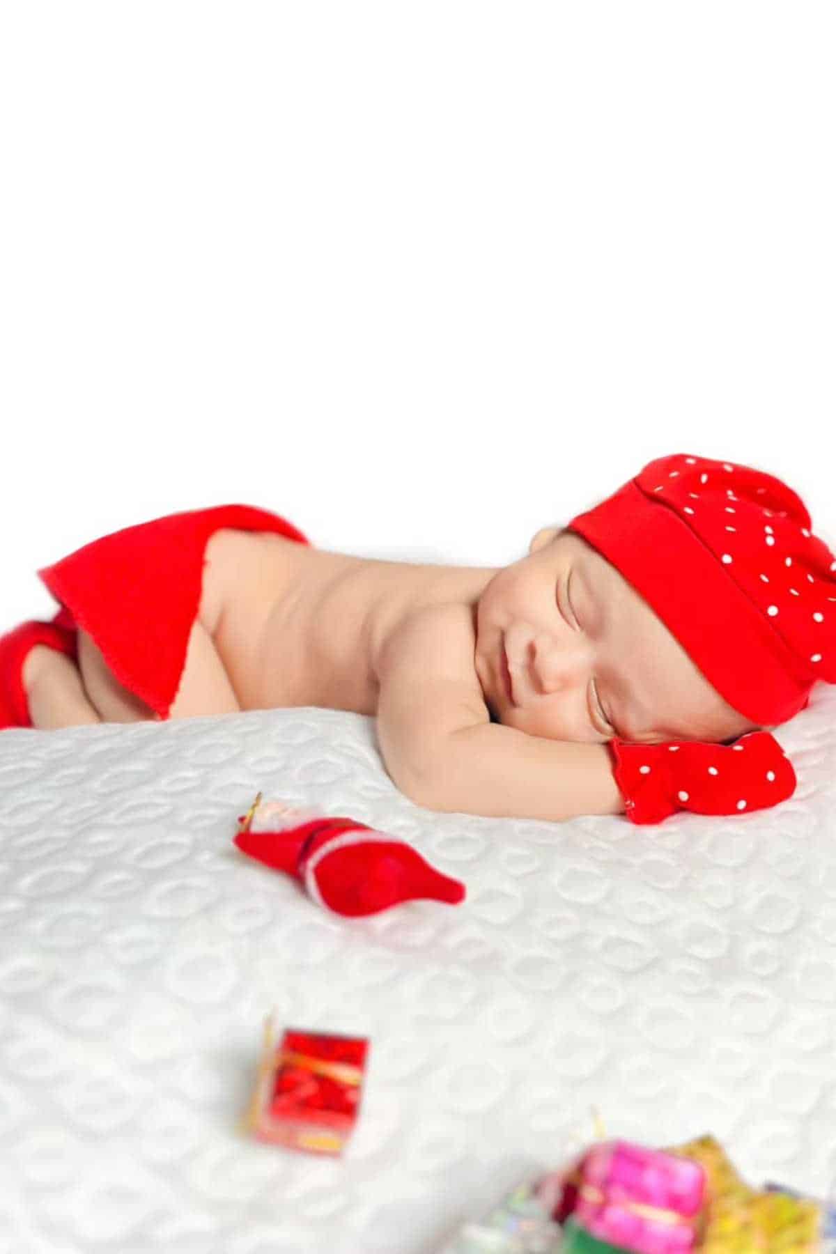 Newborn baby wearing red hat, mittens, and scarf sleeps on a white textured surface with small colorful items in the foreground.
