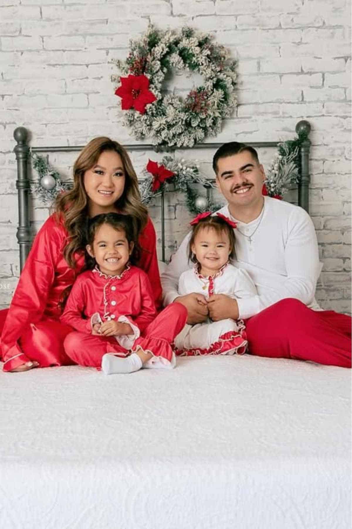 A family of four sits on a bed in festive red and white outfits, with a holiday wreath hanging on a brick wall behind them.