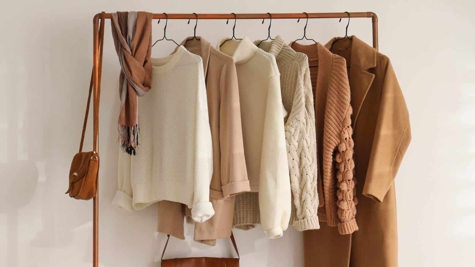 A clothing rack holds neutral-toned sweaters, a scarf, a coat, and two brown bags against a plain wall.