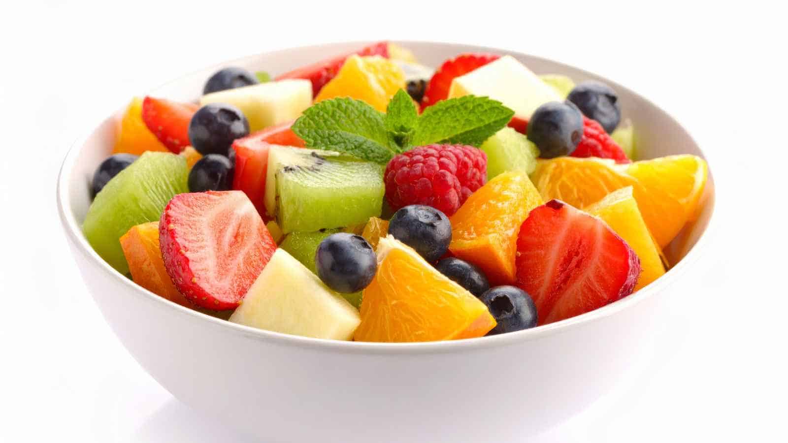 A white bowl filled with assorted fresh fruit pieces, including strawberries, kiwi, orange, apple, blueberries, and raspberries, garnished with a mint sprig.