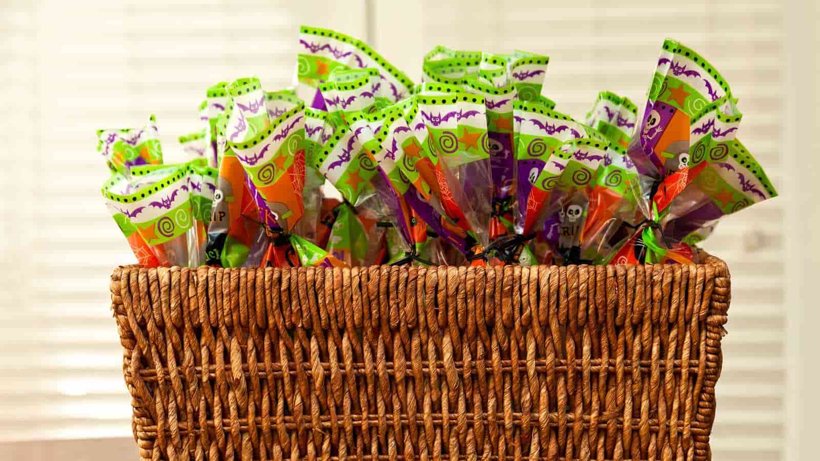 A wicker basket filled with colorful Halloween-themed treat bags, featuring green, orange, and purple packaging with festive designs.
